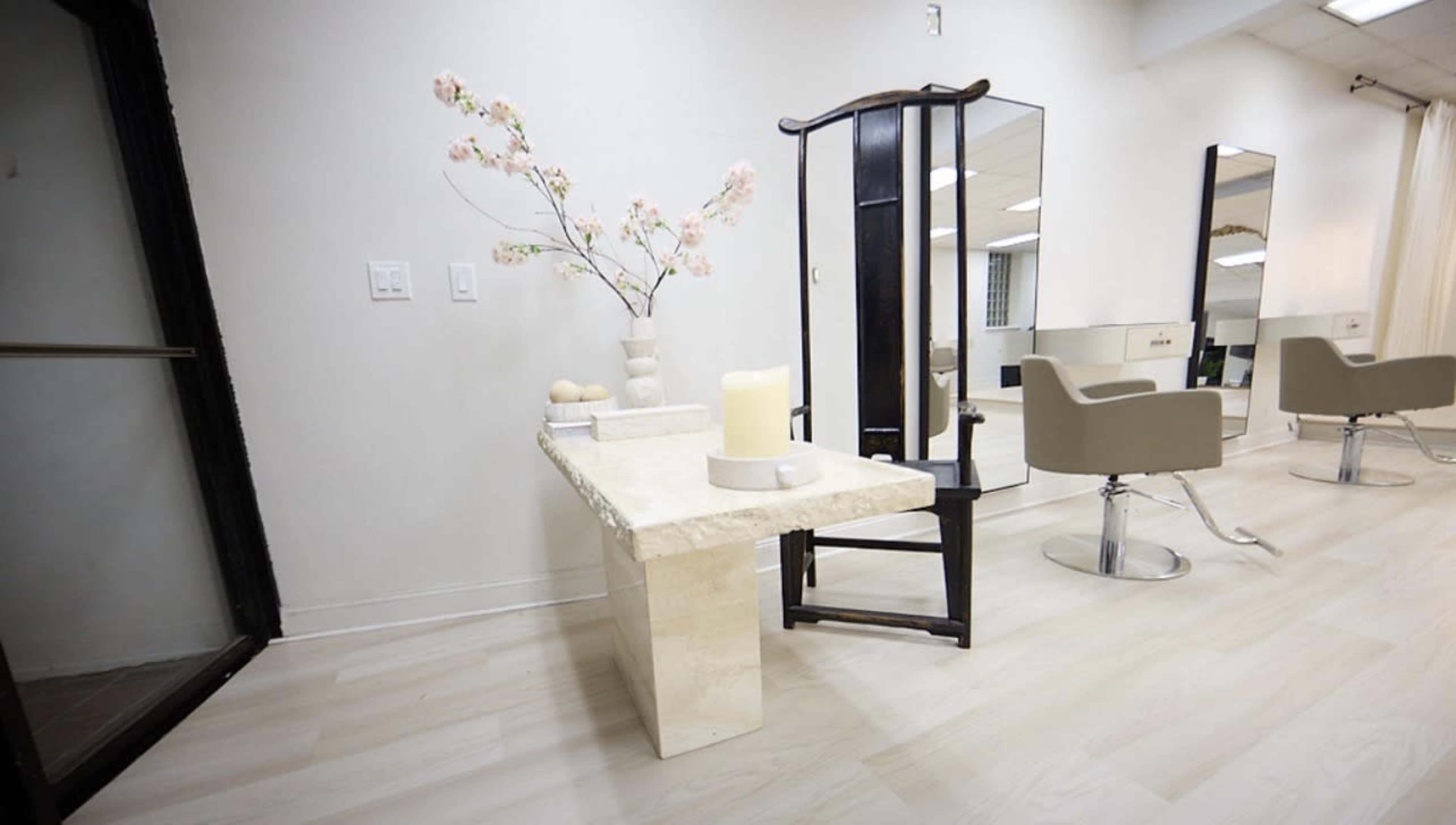 The image shows a modern hair salon interior with a minimalist stone table, a decorative vase with flowers, and sleek salon chairs arranged near mirrors.