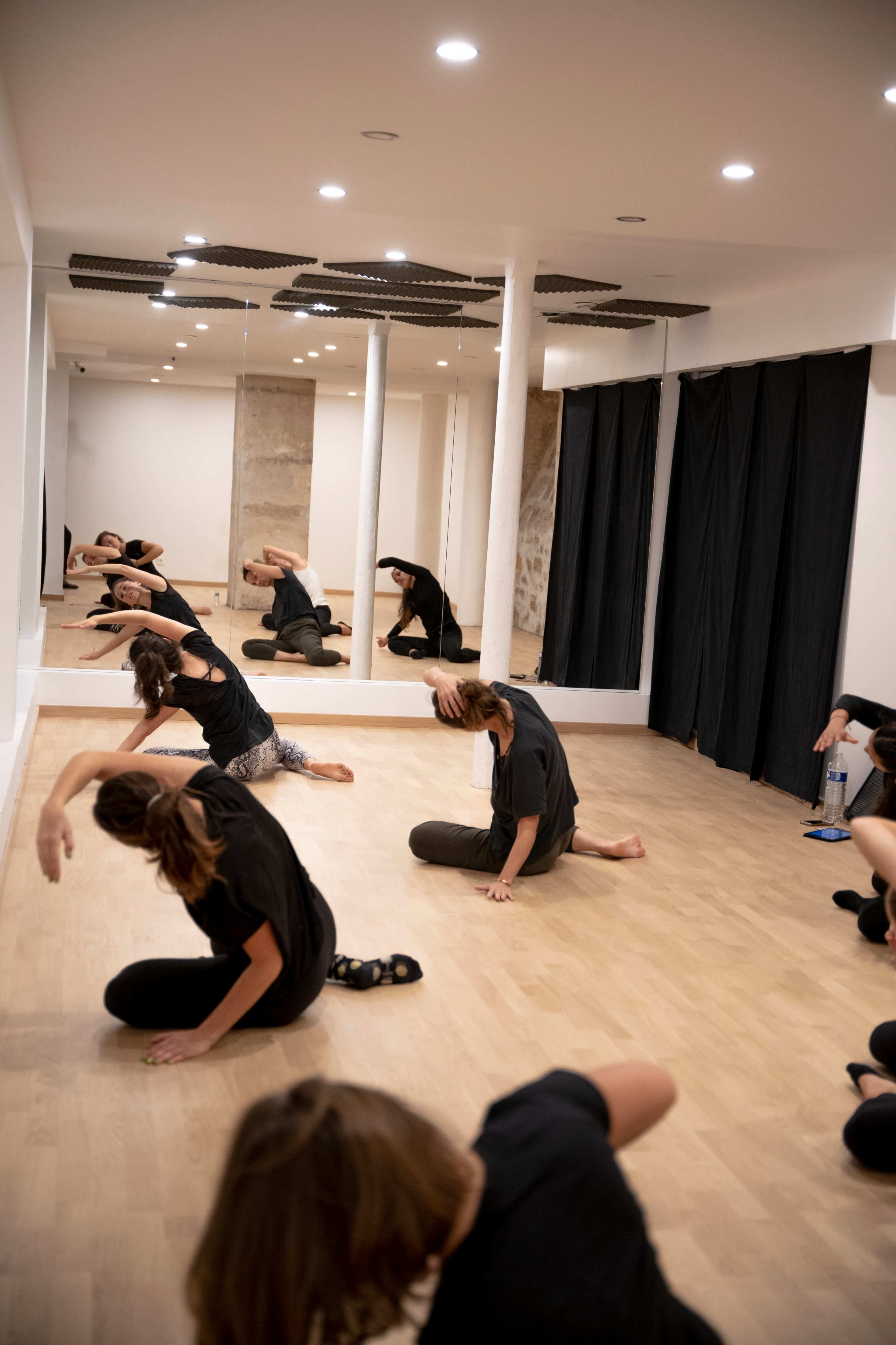 A group of people dressed in black t-shirts are performing stretching exercises in a mirrored dance studio with wooden flooring.