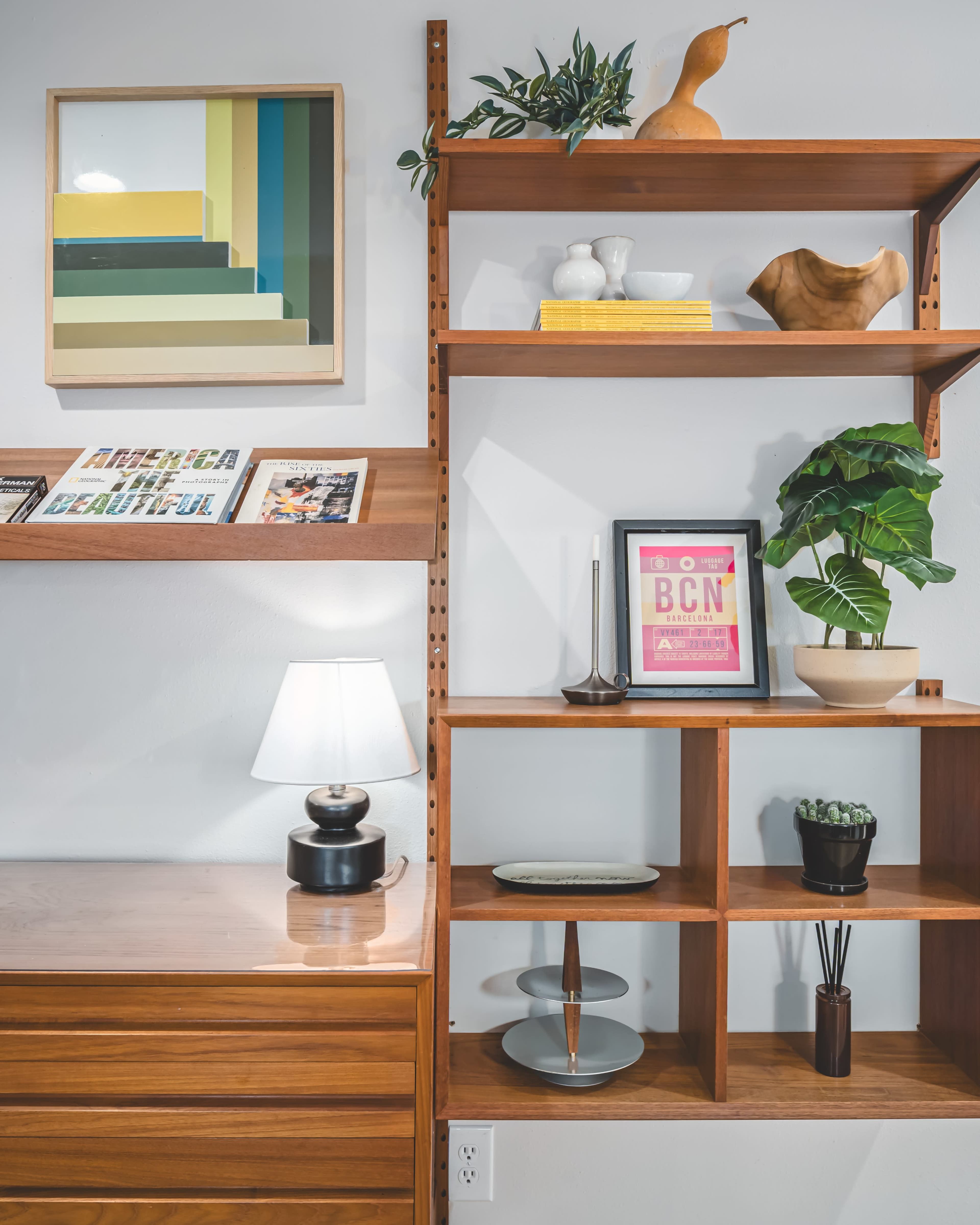 The image shows a wooden shelving unit displaying various decorative items, including plants, a lamp, and framed artwork.