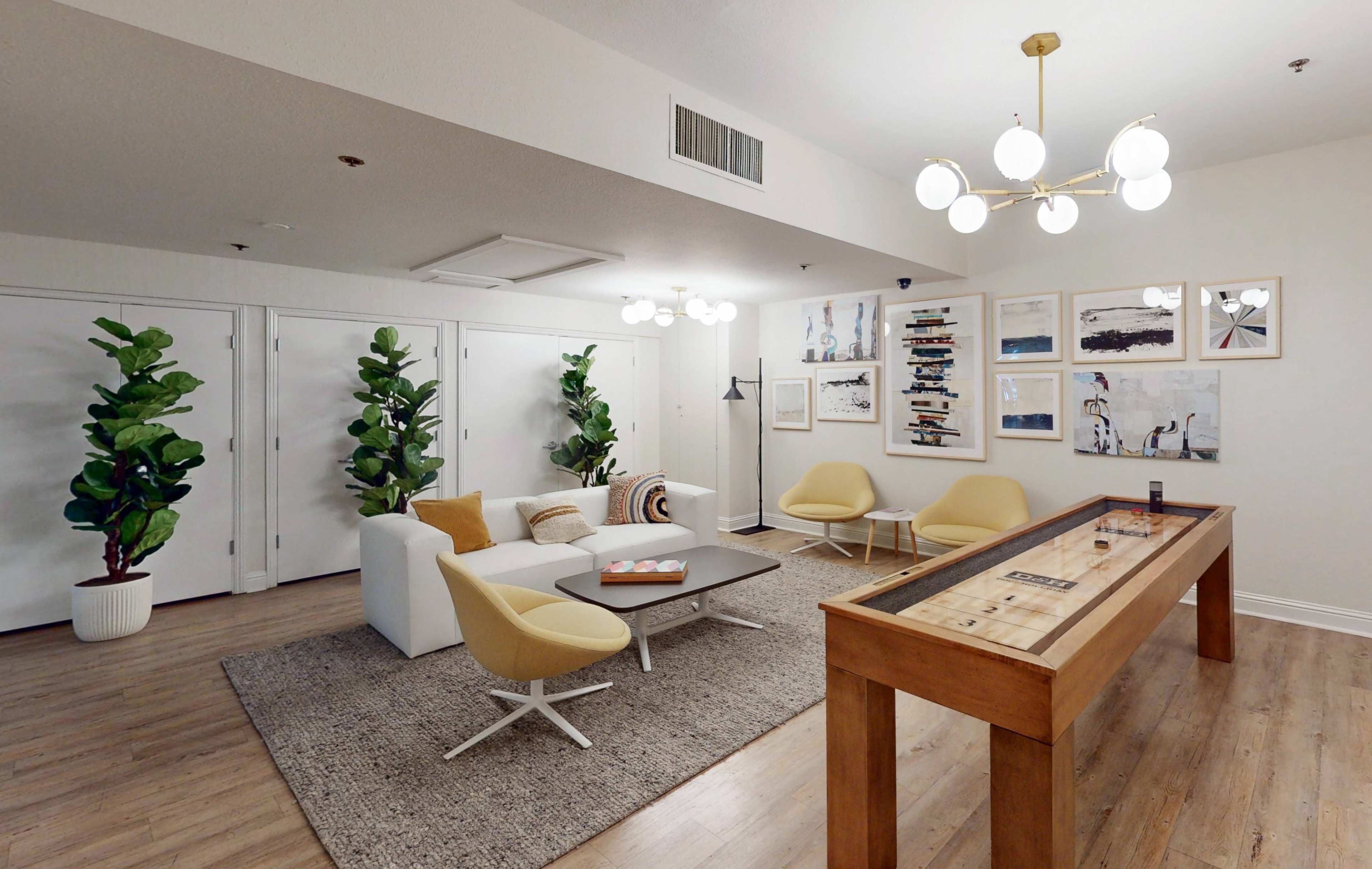 The image shows a modern lounge area featuring a white sectional sofa, yellow accent chairs, a wooden game table, and framed artwork hanging on the walls.