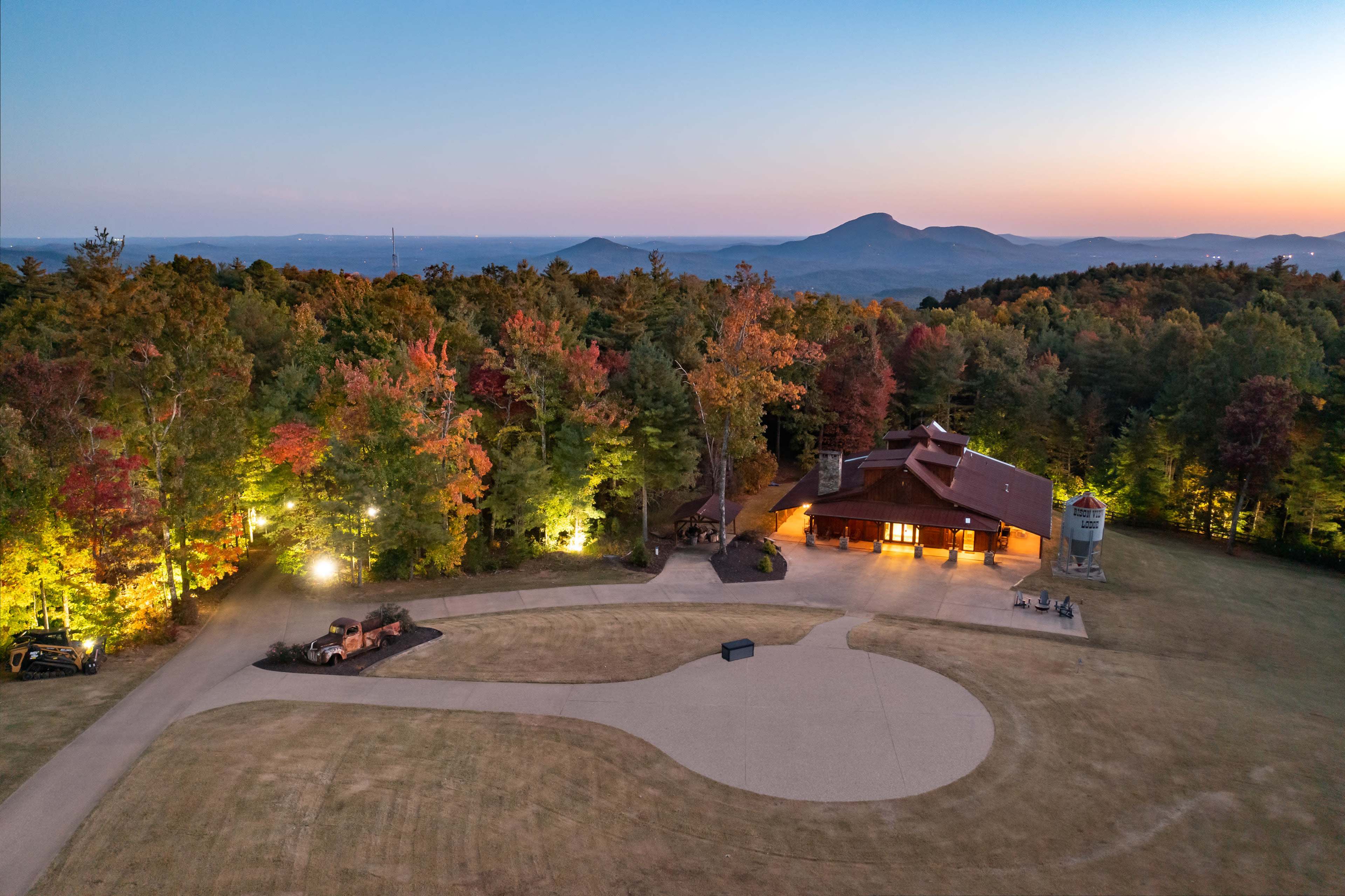 The image shows a spacious lodge surrounded by trees with autumn foliage and a mountain range in the background, illuminated by evening light.