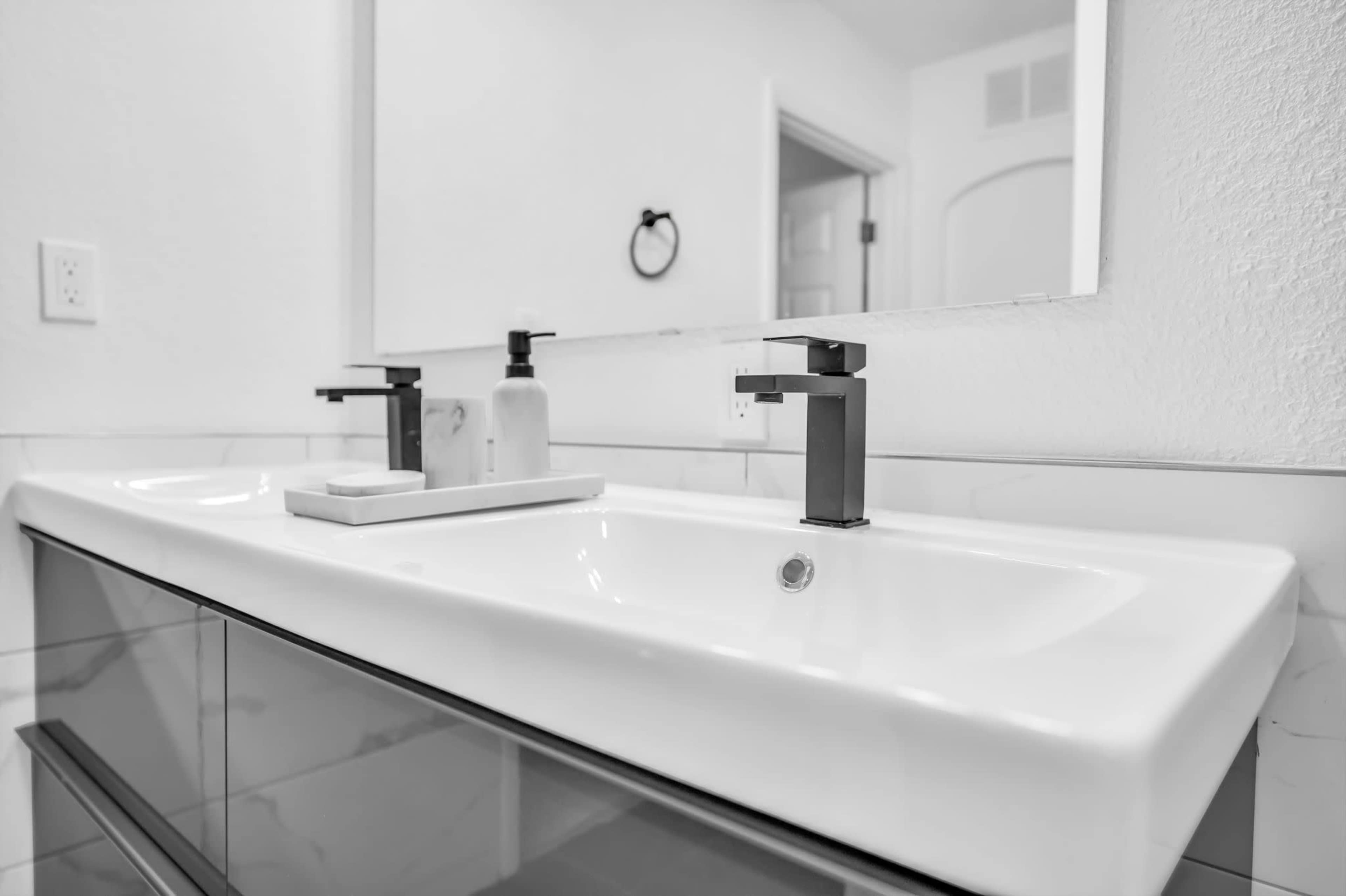 A modern bathroom sink with a sleek faucet and a small soap tray sits against a white wall, reflected in a mirror.