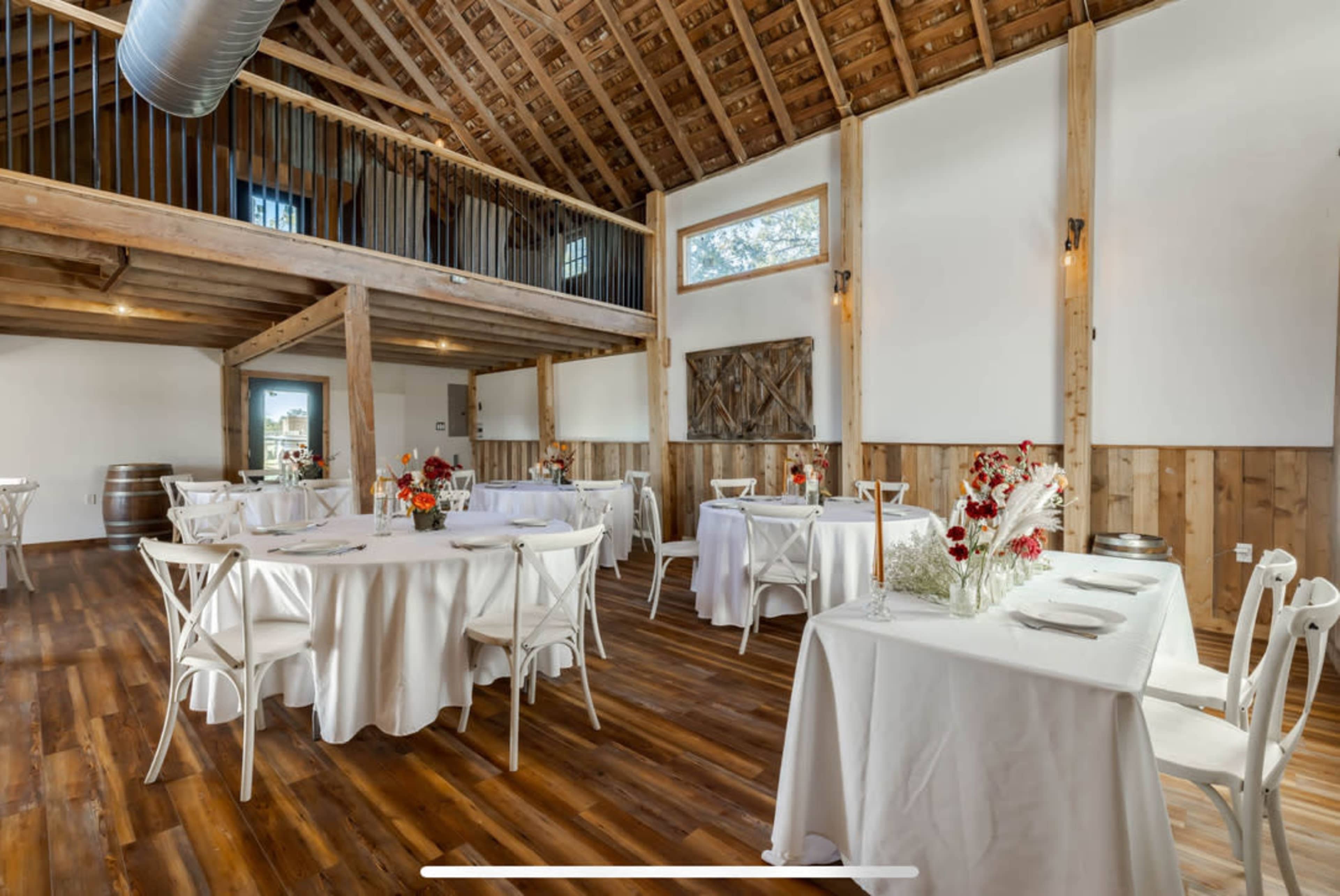 An event space with several round tables covered in white tablecloths, surrounded by wooden beams and walls, with large windows letting in natural light.