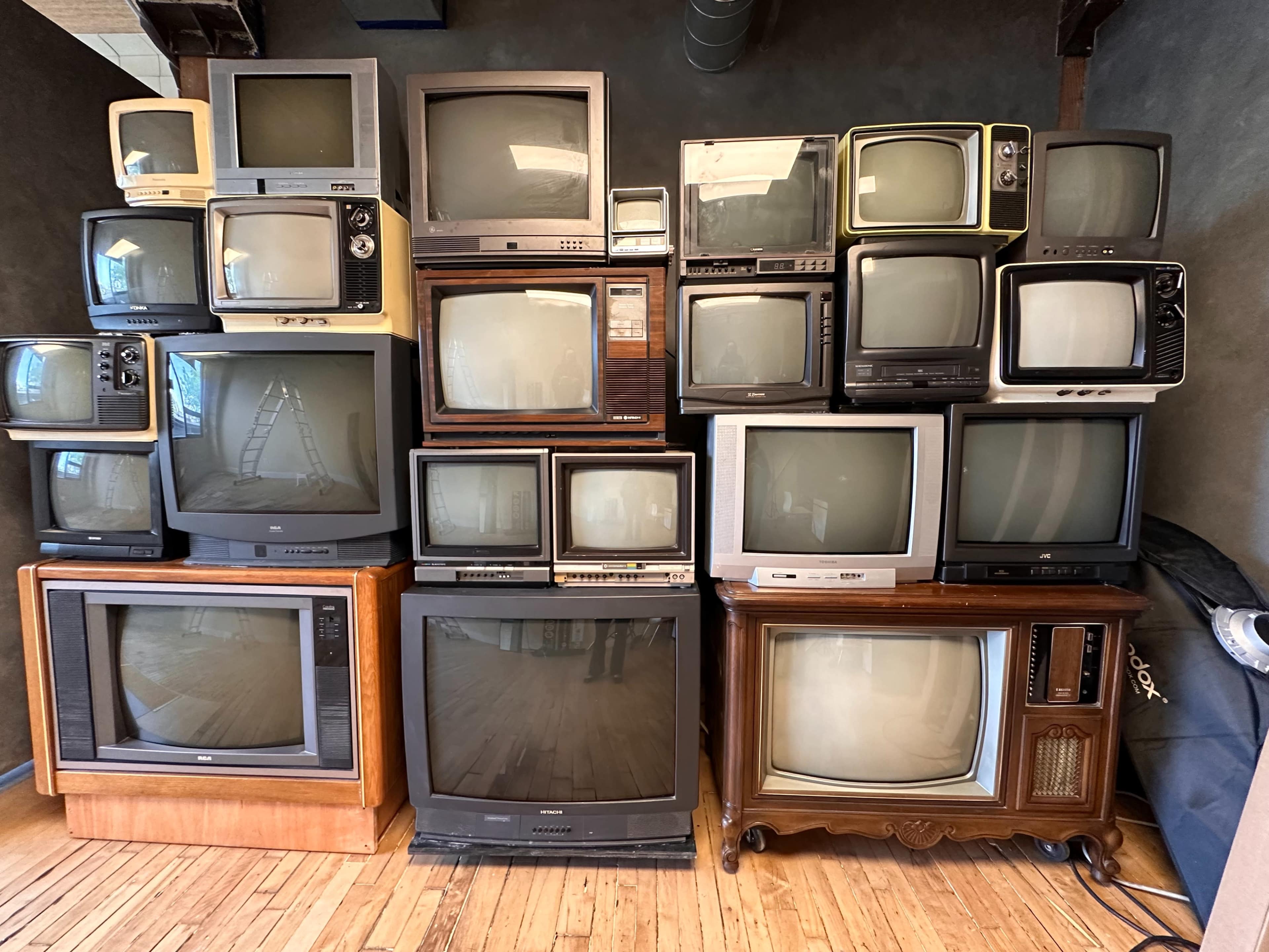 A stack of vintage televisions in varying sizes and styles is arranged against a wall in a room with wooden floors.