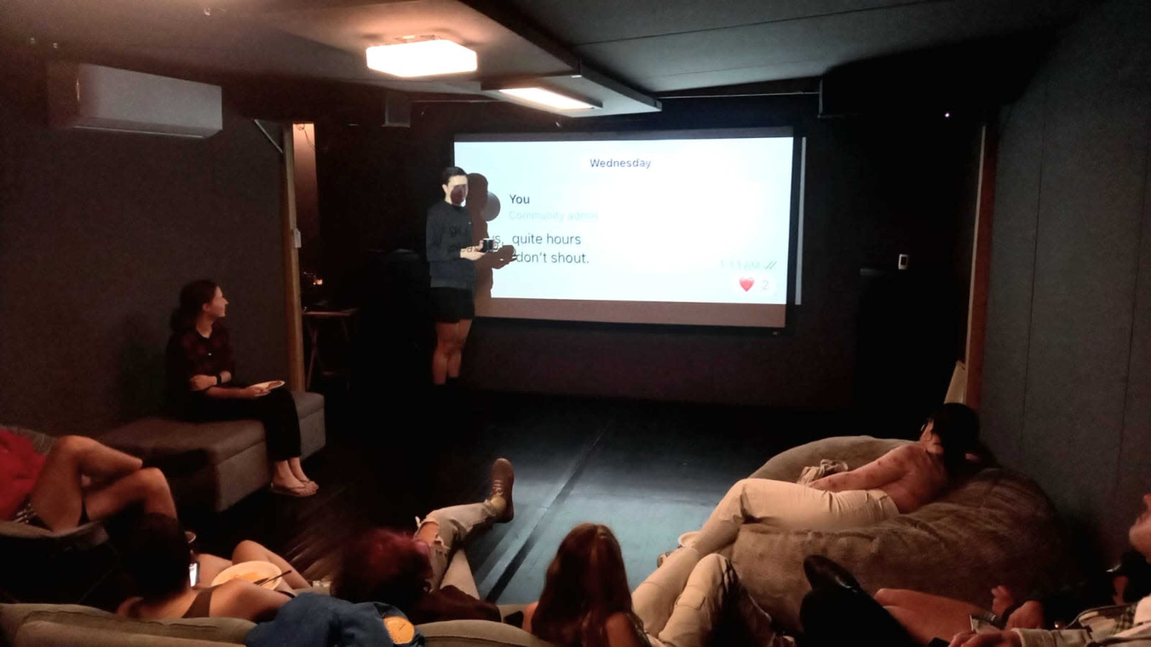 A group of people sits on bean bags in a dimly lit room while one person presents information on a screen.
