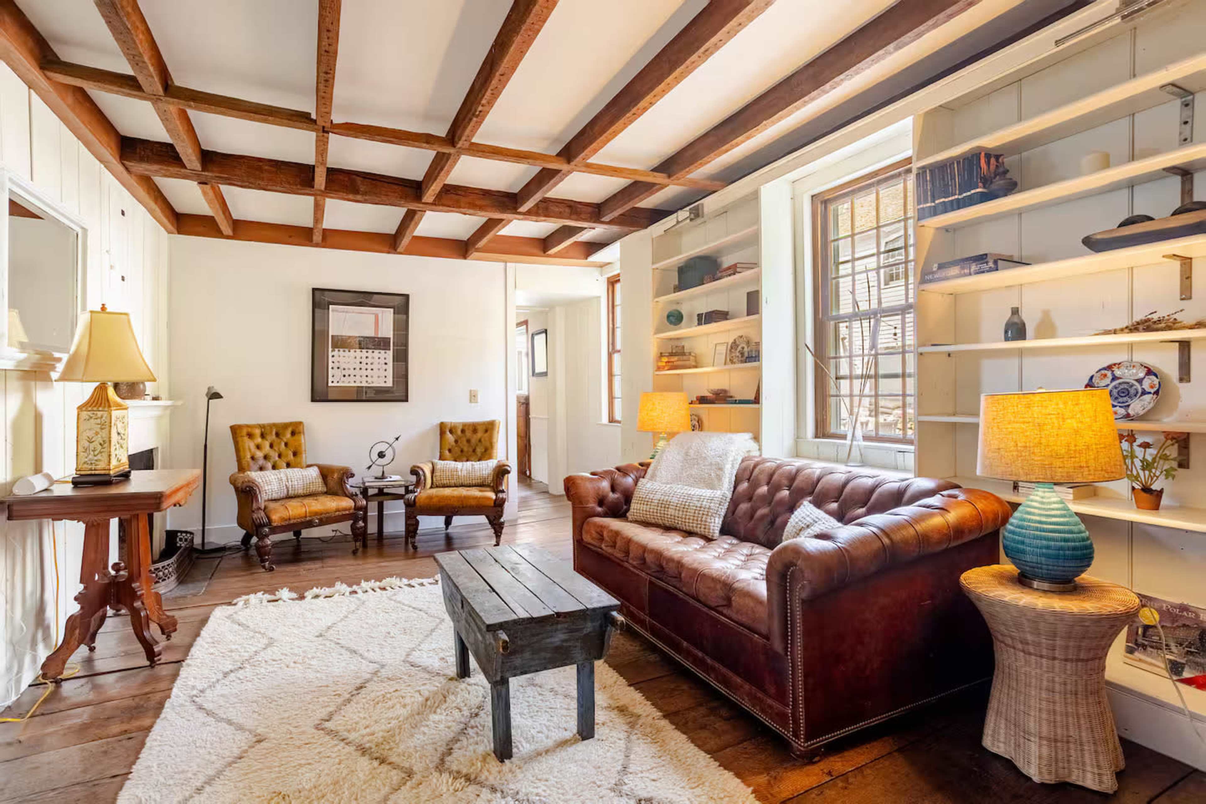 A cozy living room features a brown leather chesterfield sofa, two upholstered armchairs, a wooden coffee table, and shelves filled with decorative items.
