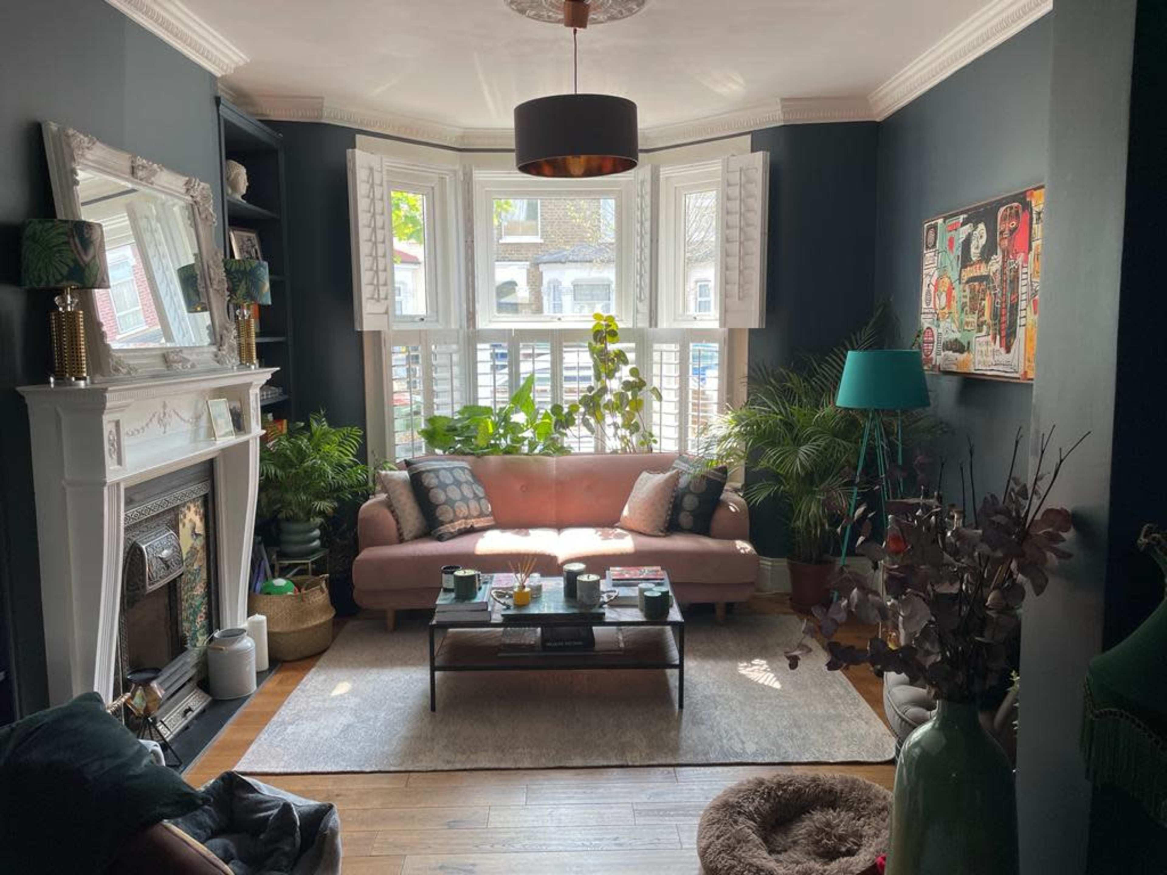 A stylish living room features a pink sofa, a coffee table, and several indoor plants, with large bay windows allowing natural light to enter.