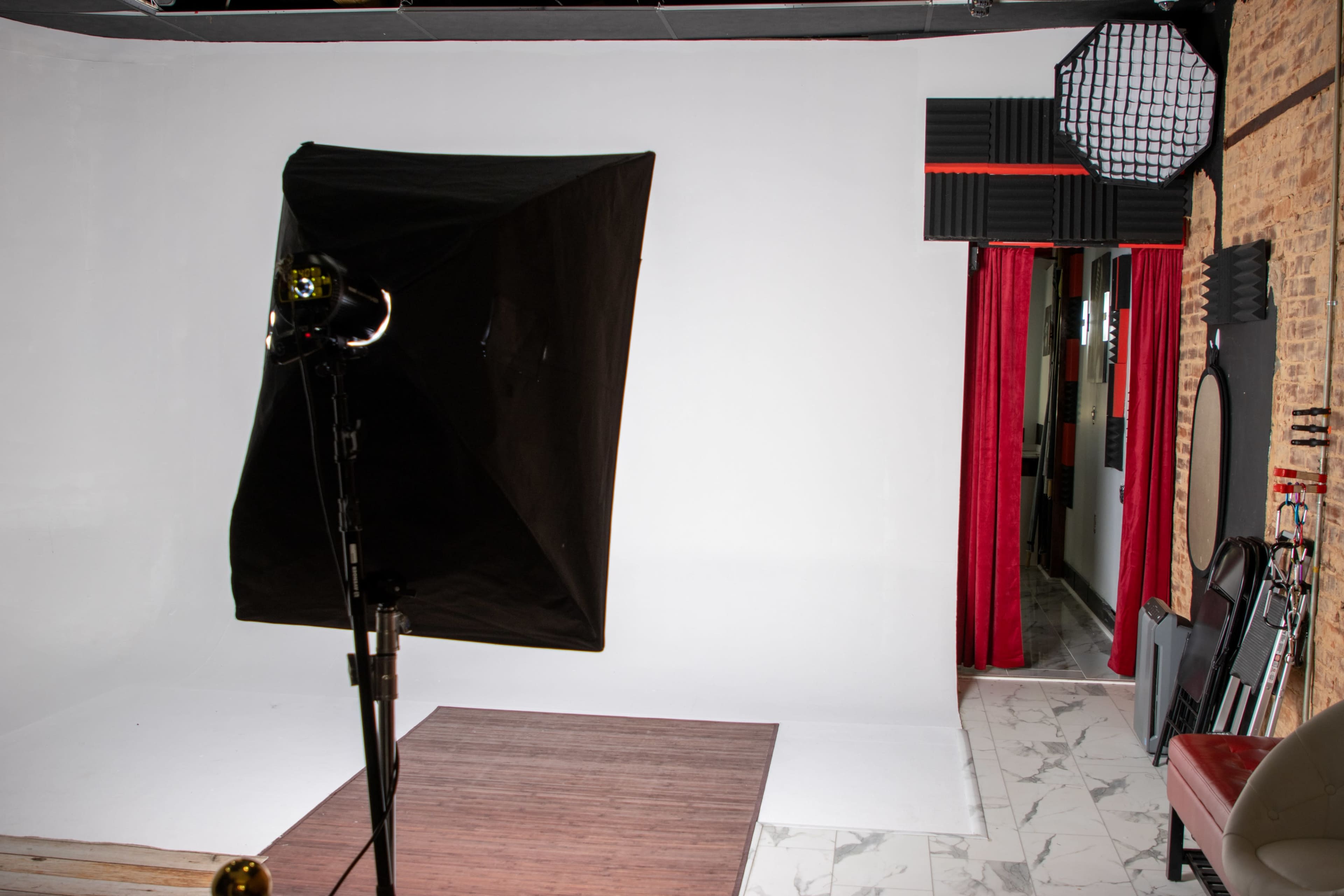 A photography studio with a softbox light, a white backdrop, and a red curtain leading to another area.