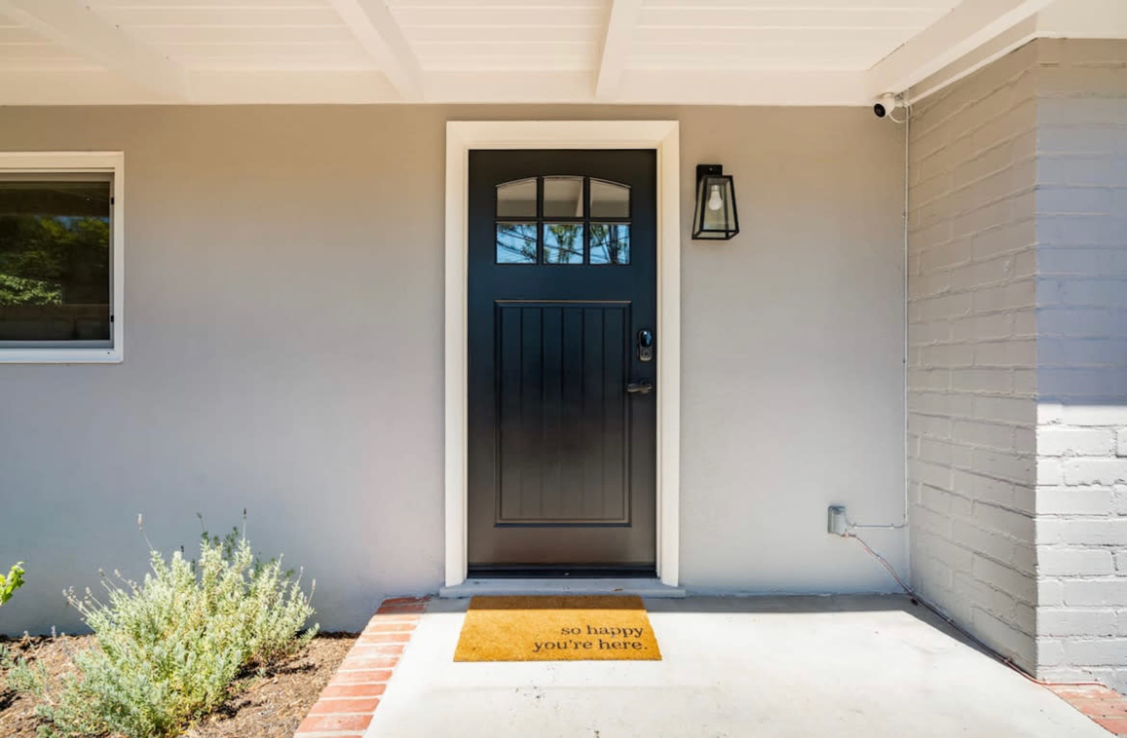 A front door with a dark wooden finish and a welcome mat that reads "so happy you're here," set in a modern, light-colored house exterior.