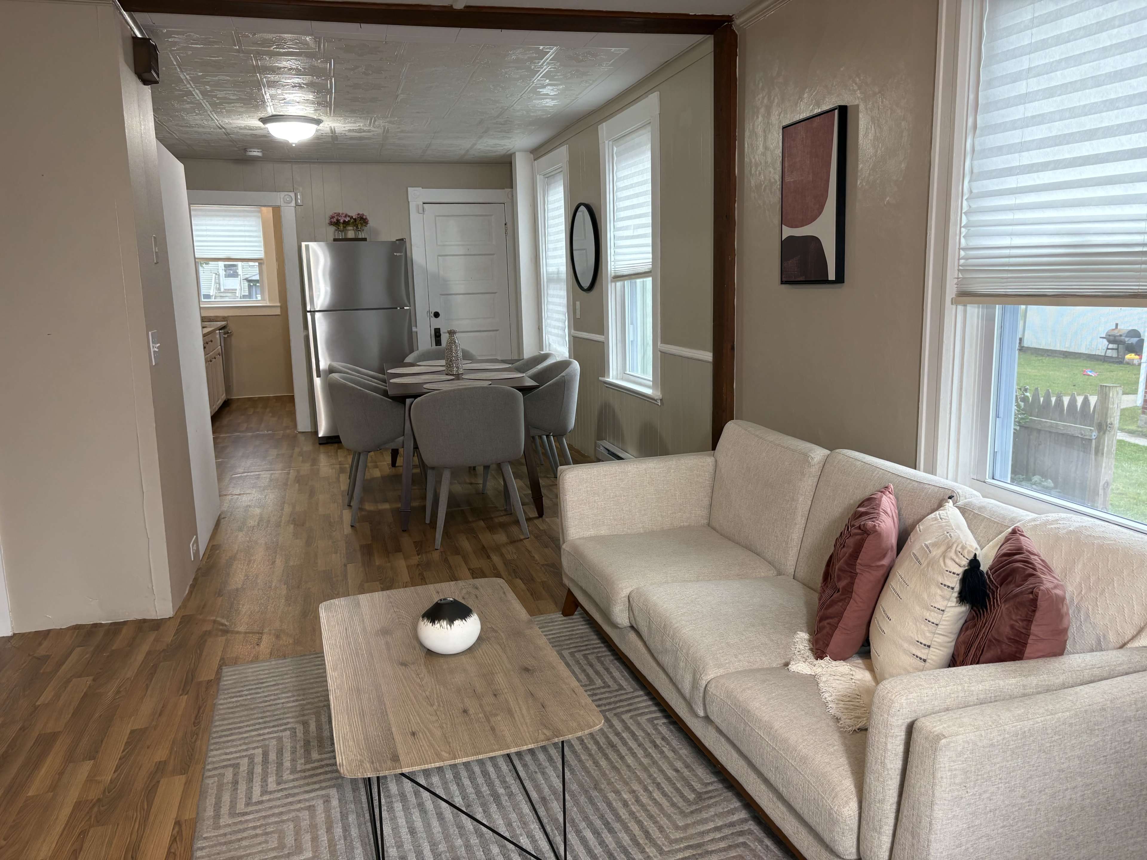 The image shows a modern living space featuring a light-colored sofa, a coffee table, and a dining area with a round table and four chairs, all set in a room with wooden flooring and large windows.