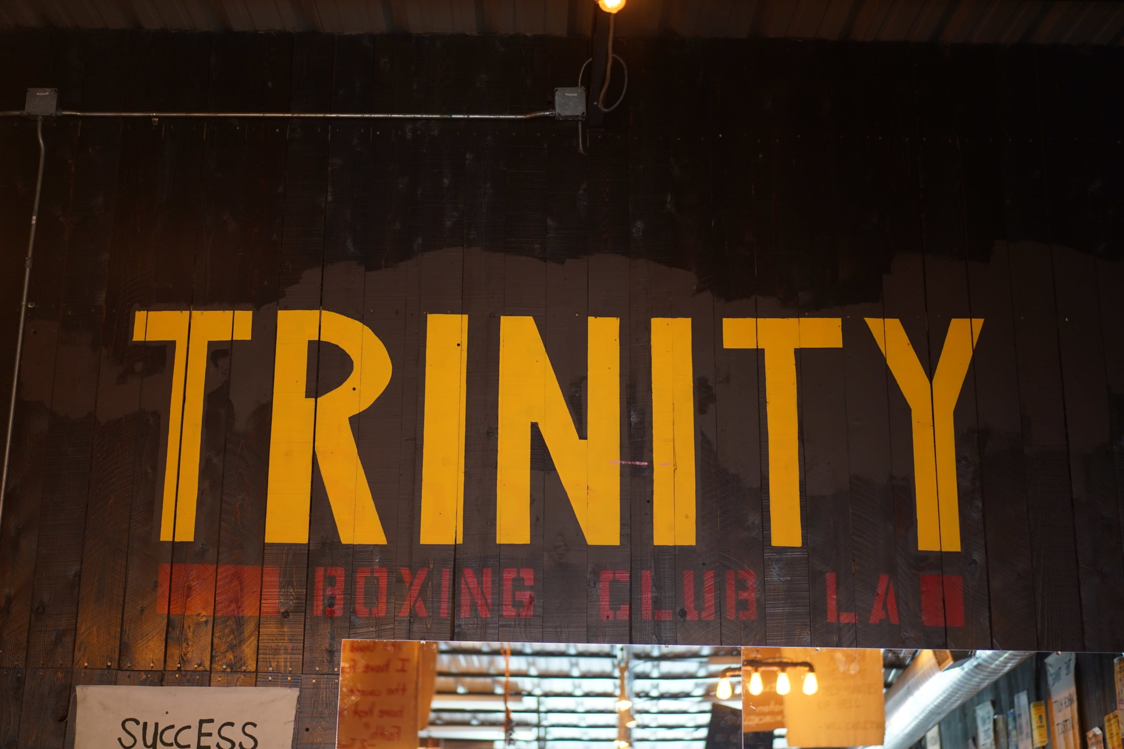 A large sign that reads "TRINITY BOXING CLUB LA" in bold yellow and red letters on a dark wooden background.