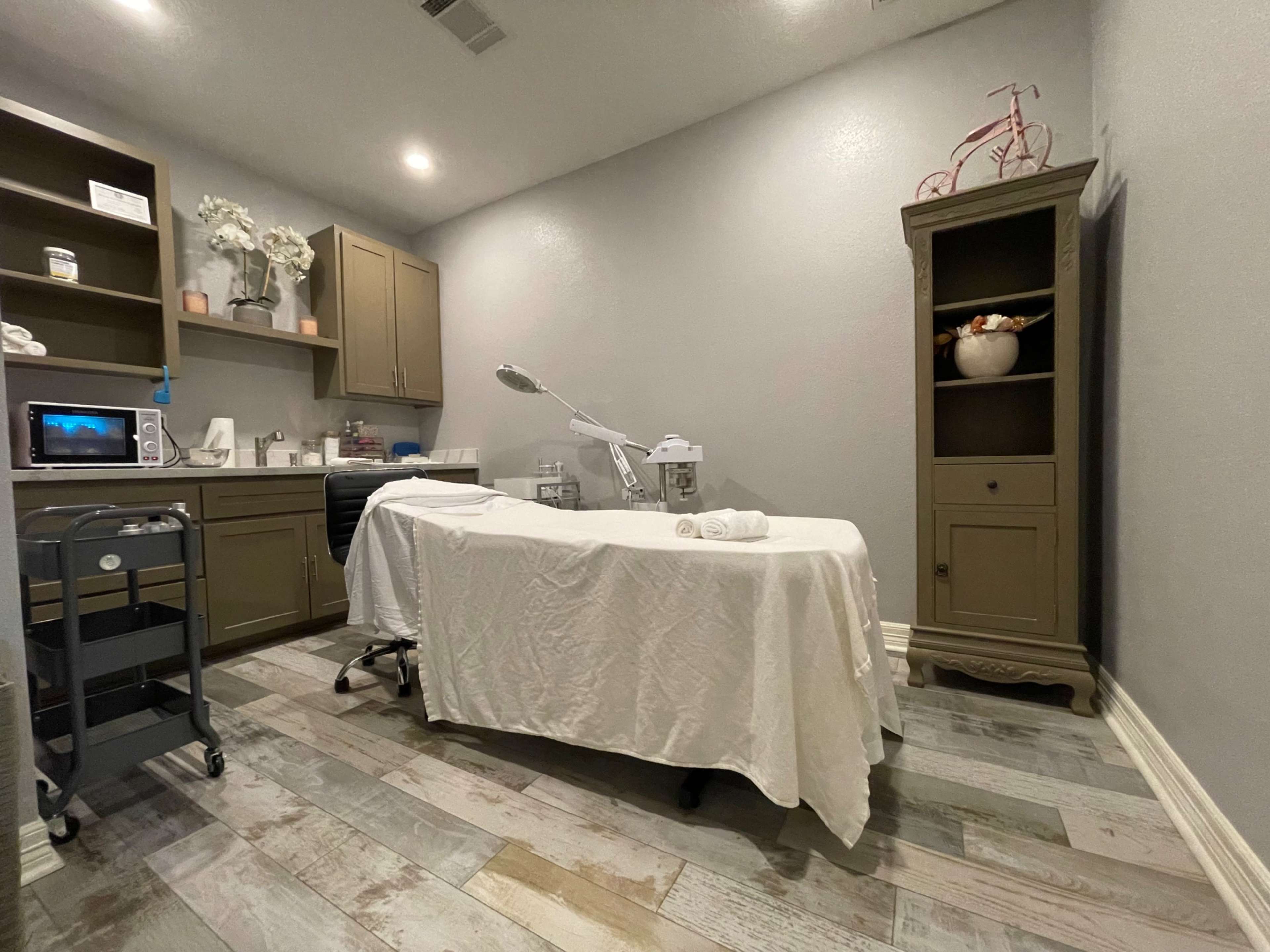 A treatment room with a covered massage table, a chair, cabinets, and a small shelf with decorative items.