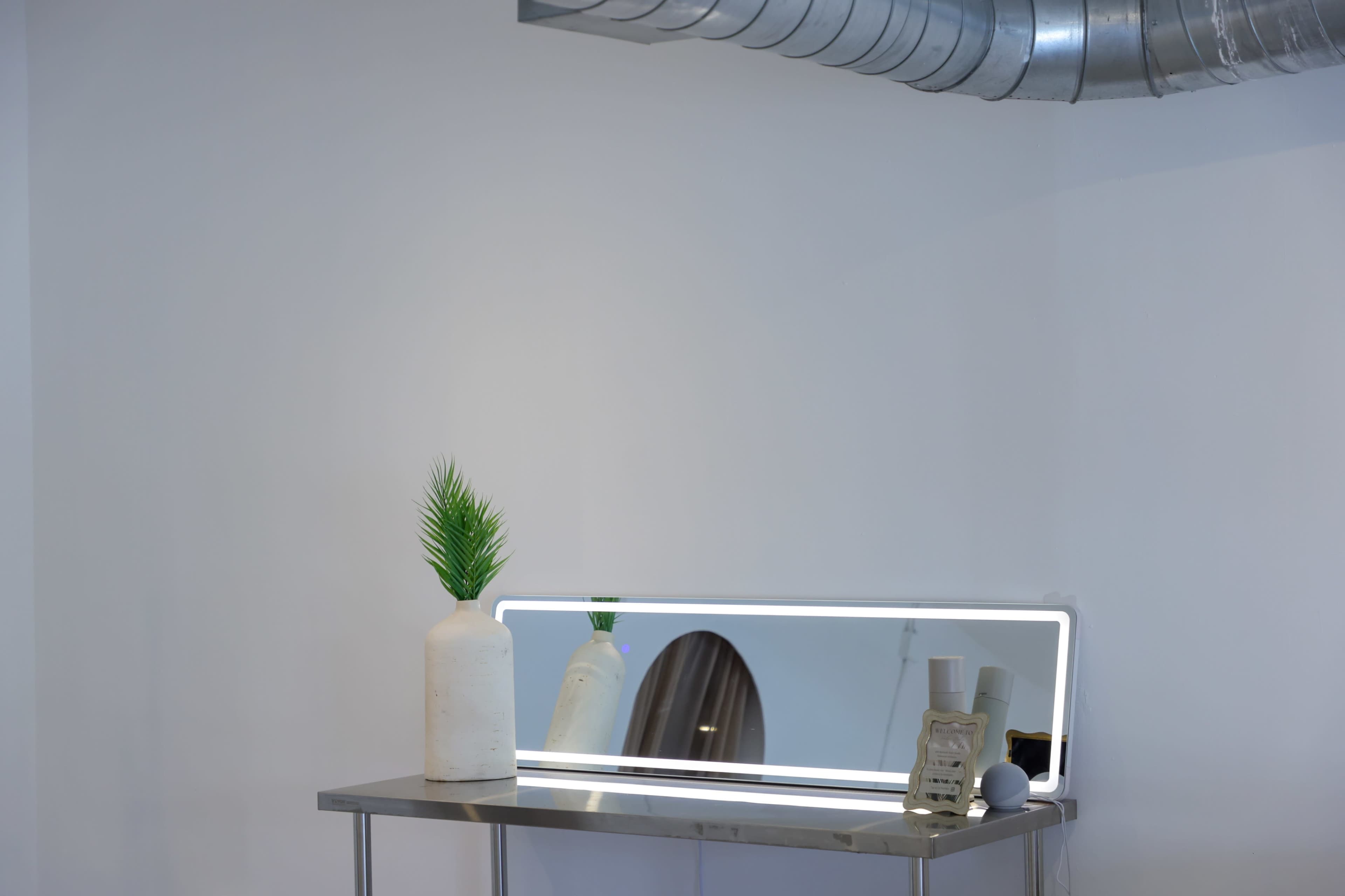 A sleek vanity table with a mirrored surface, a modern vase containing a faux plant, and a vintage telephone is positioned against a white wall with exposed ductwork above.