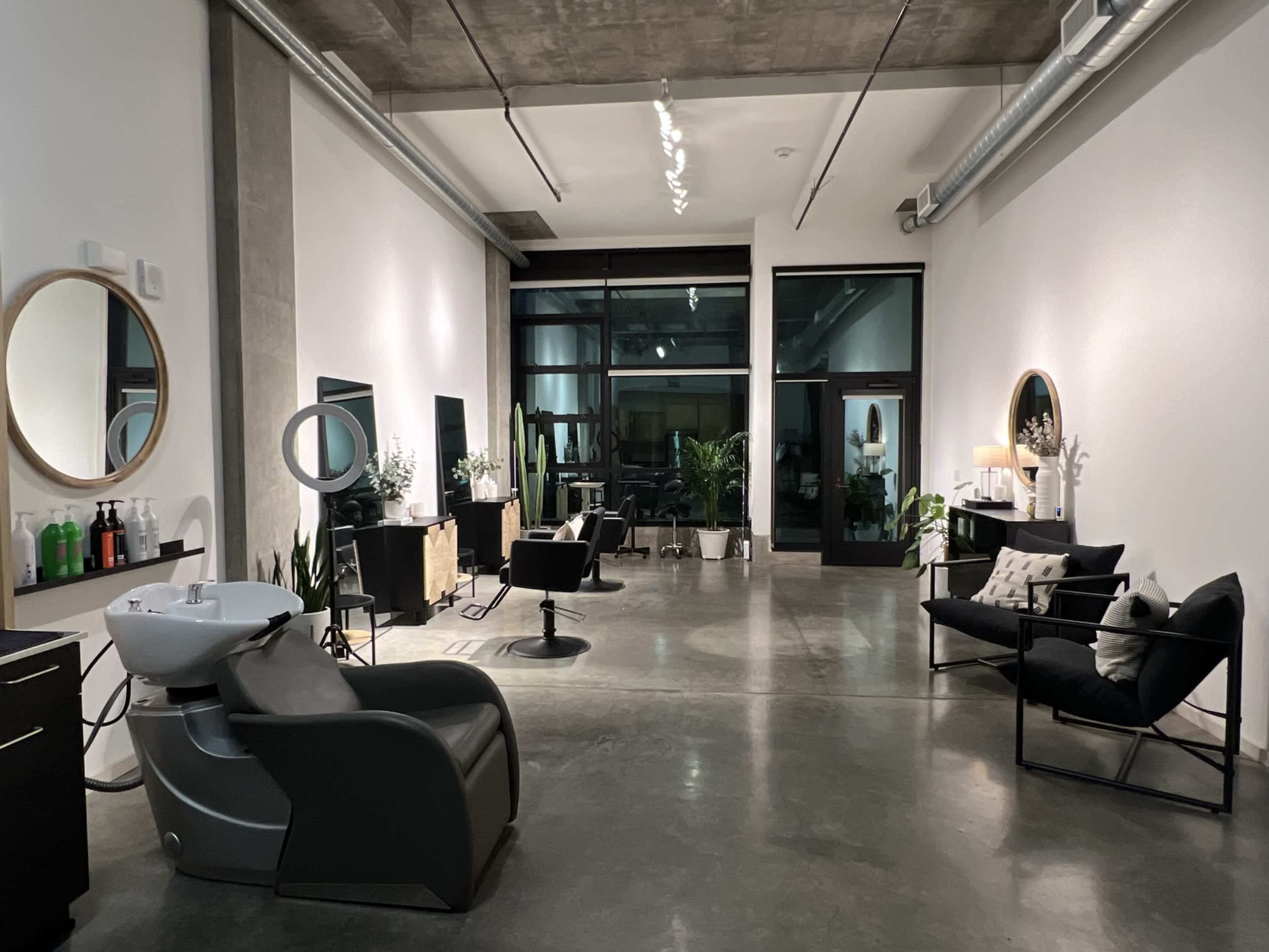A modern hair salon with several styling stations, a shampoo area, and large windows allowing natural light.