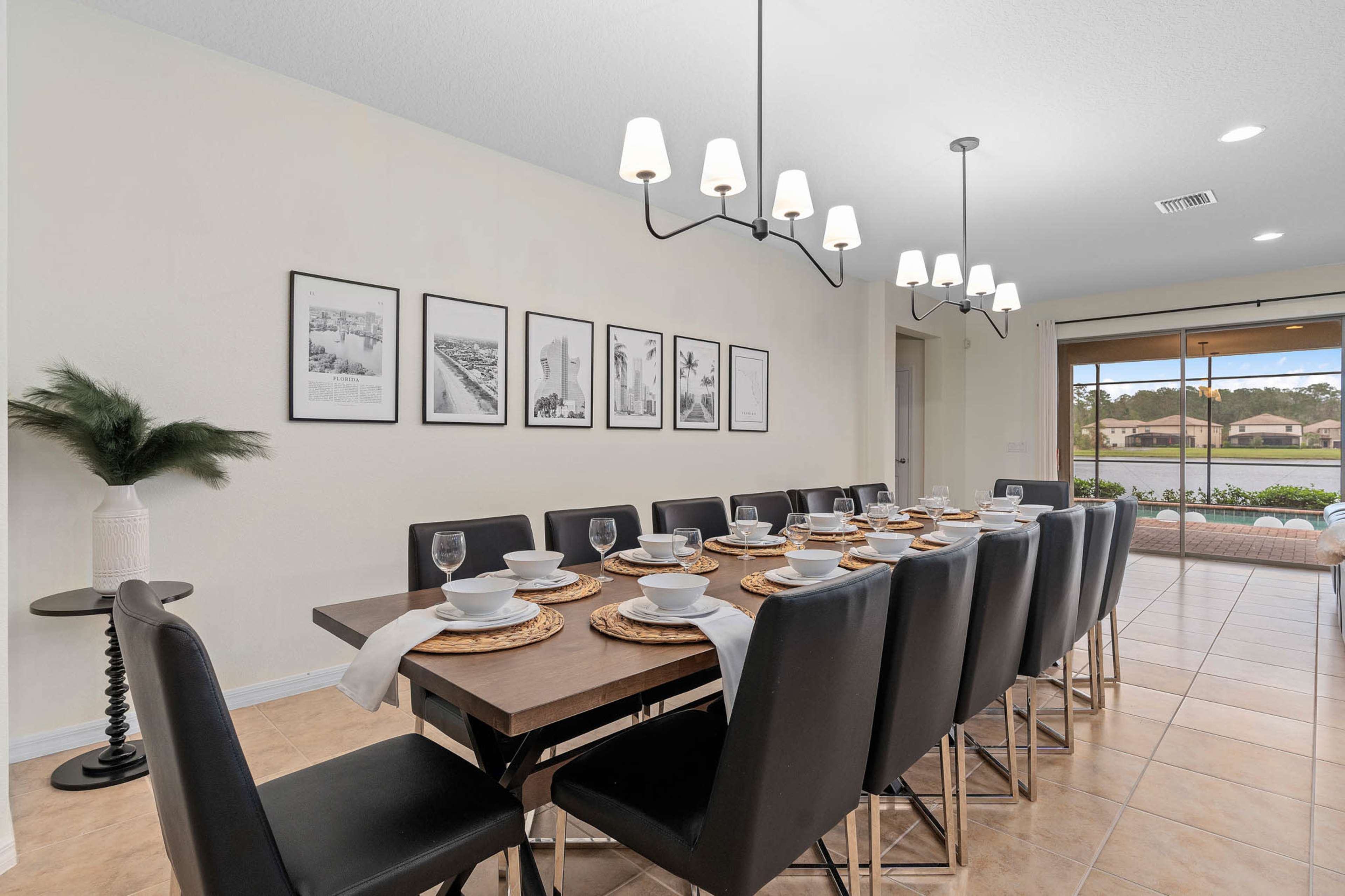 A long dining table set for a meal is displayed in a bright dining area with framed black-and-white photos on the wall and sliding glass doors leading to an outdoor space.
