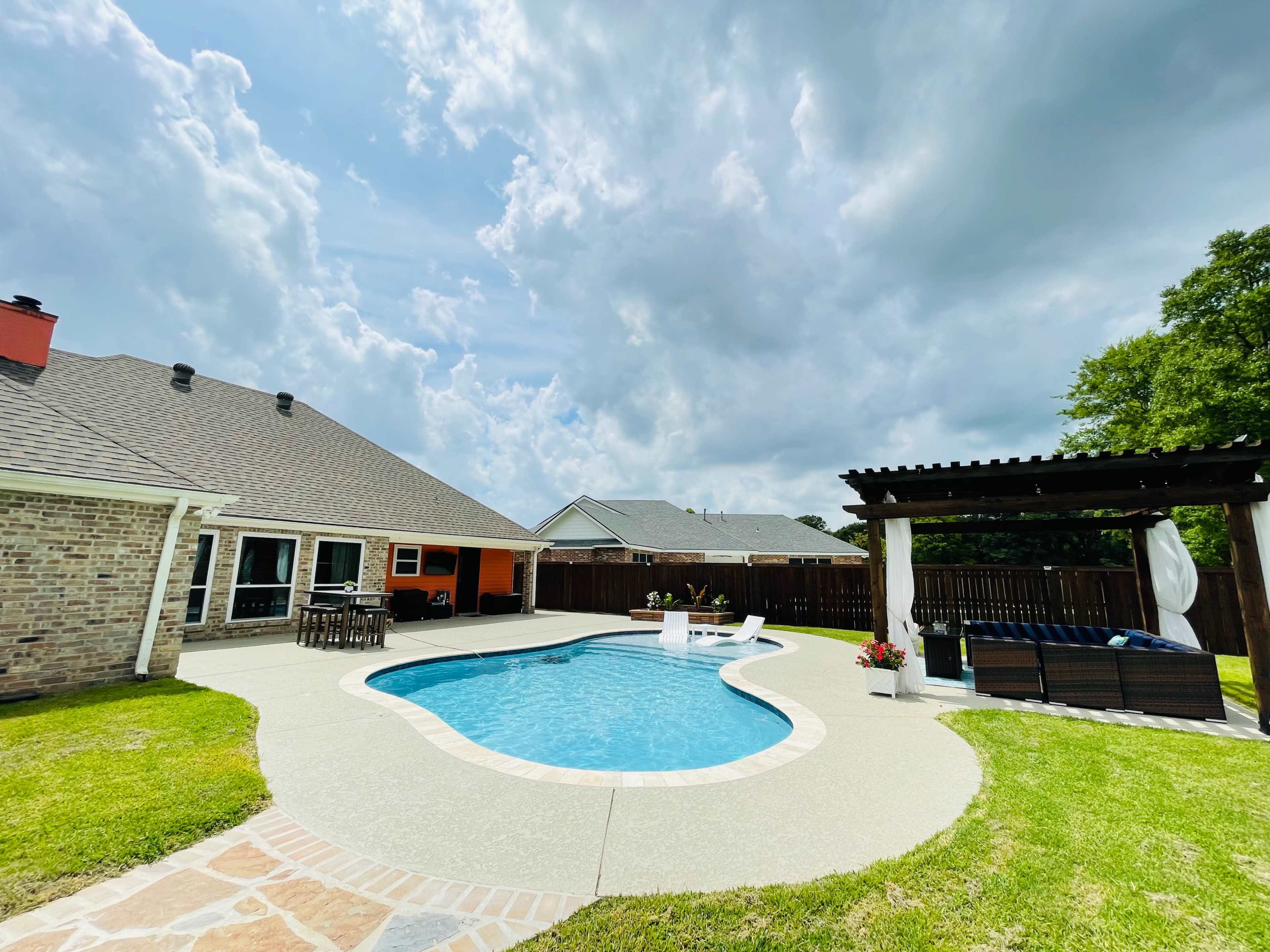 A backyard features a swimming pool with a surrounding patio, adjacent to a house and a pergola with seating.