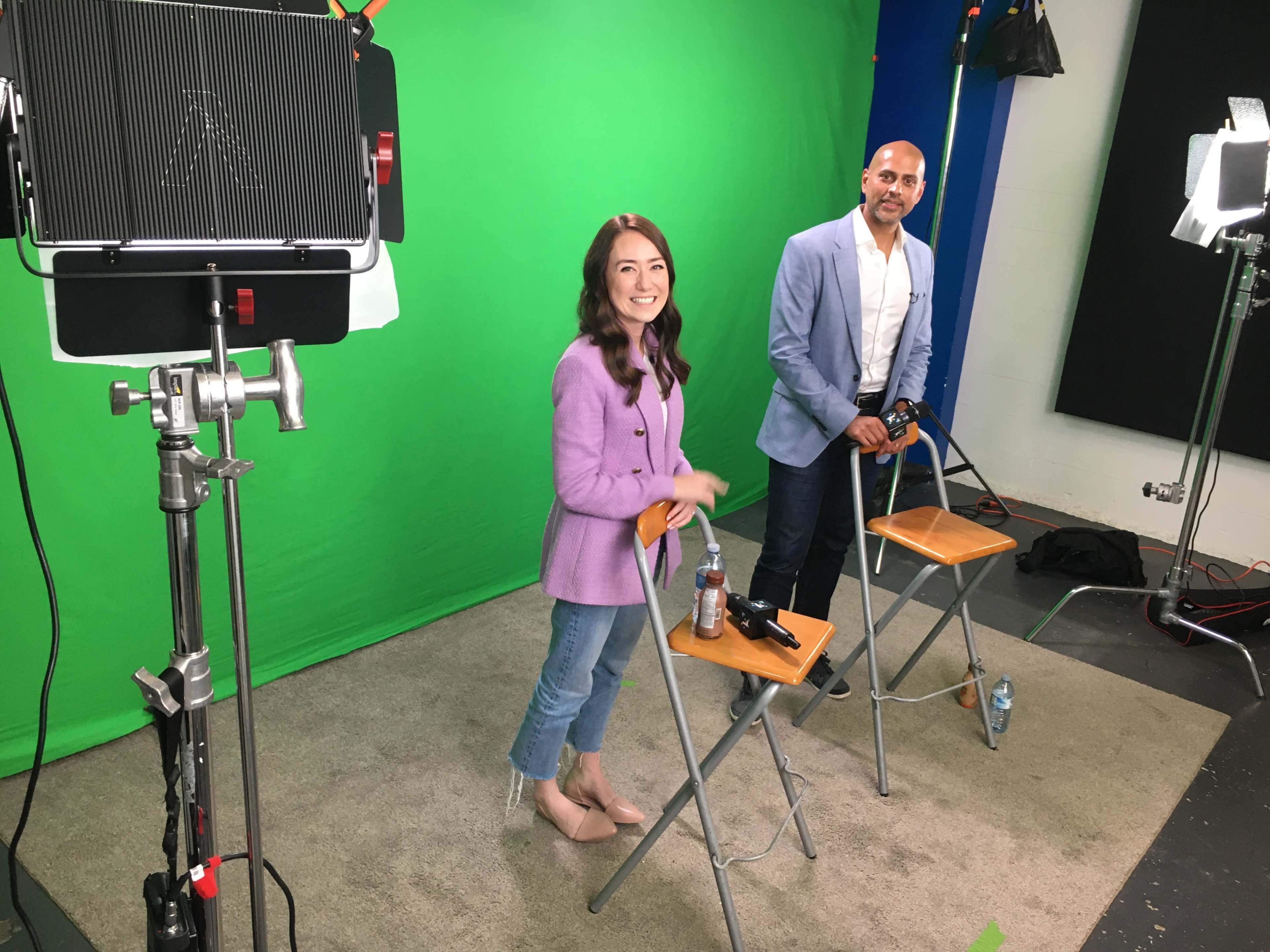 Two individuals stand on a green screen set, one in a purple blazer and the other in a light blue jacket, with chairs and equipment visible around them.