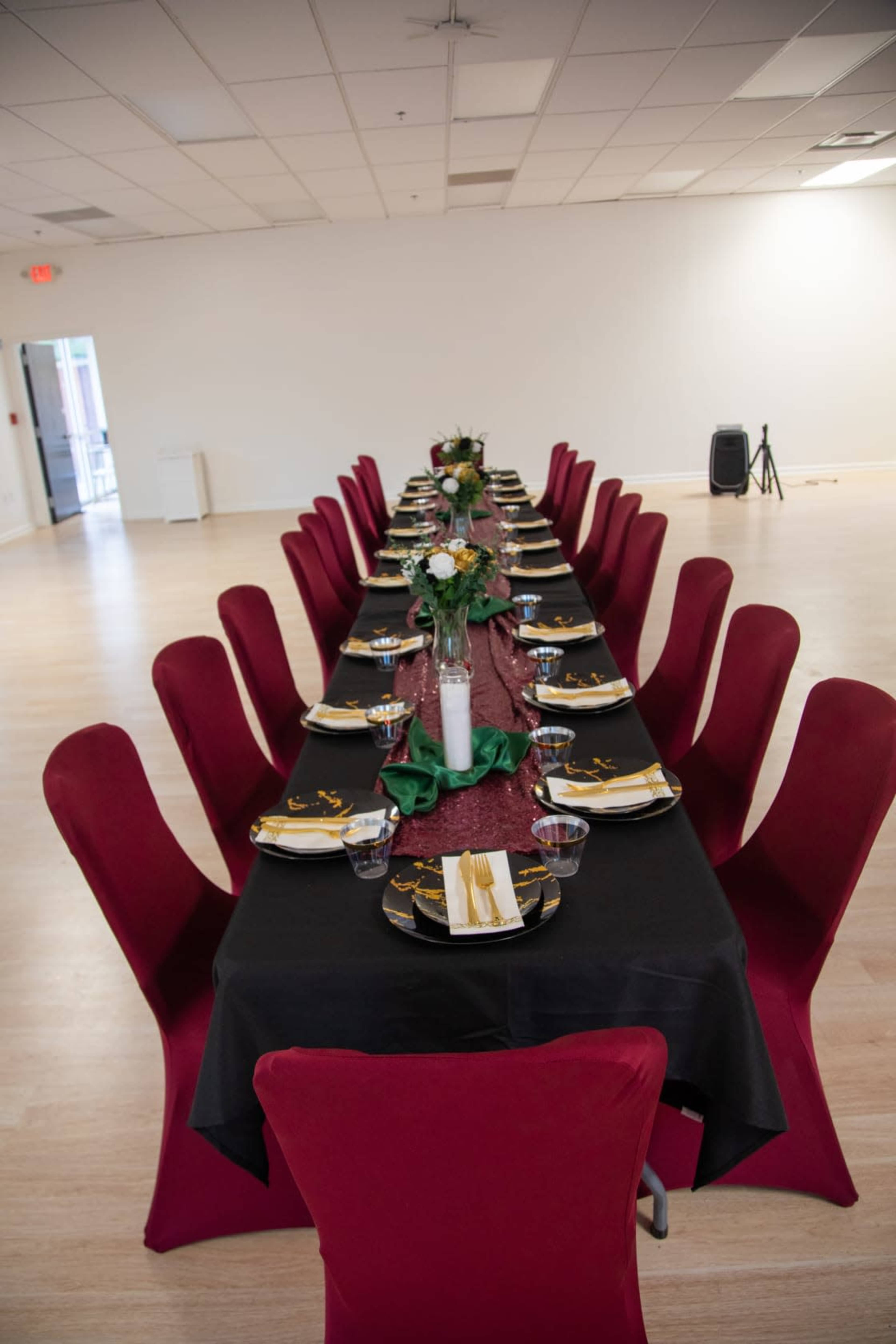 A long, elegantly set dining table is arranged with black and gold tableware, adorned with centerpieces of flowers, and surrounded by red chairs in a spacious room.