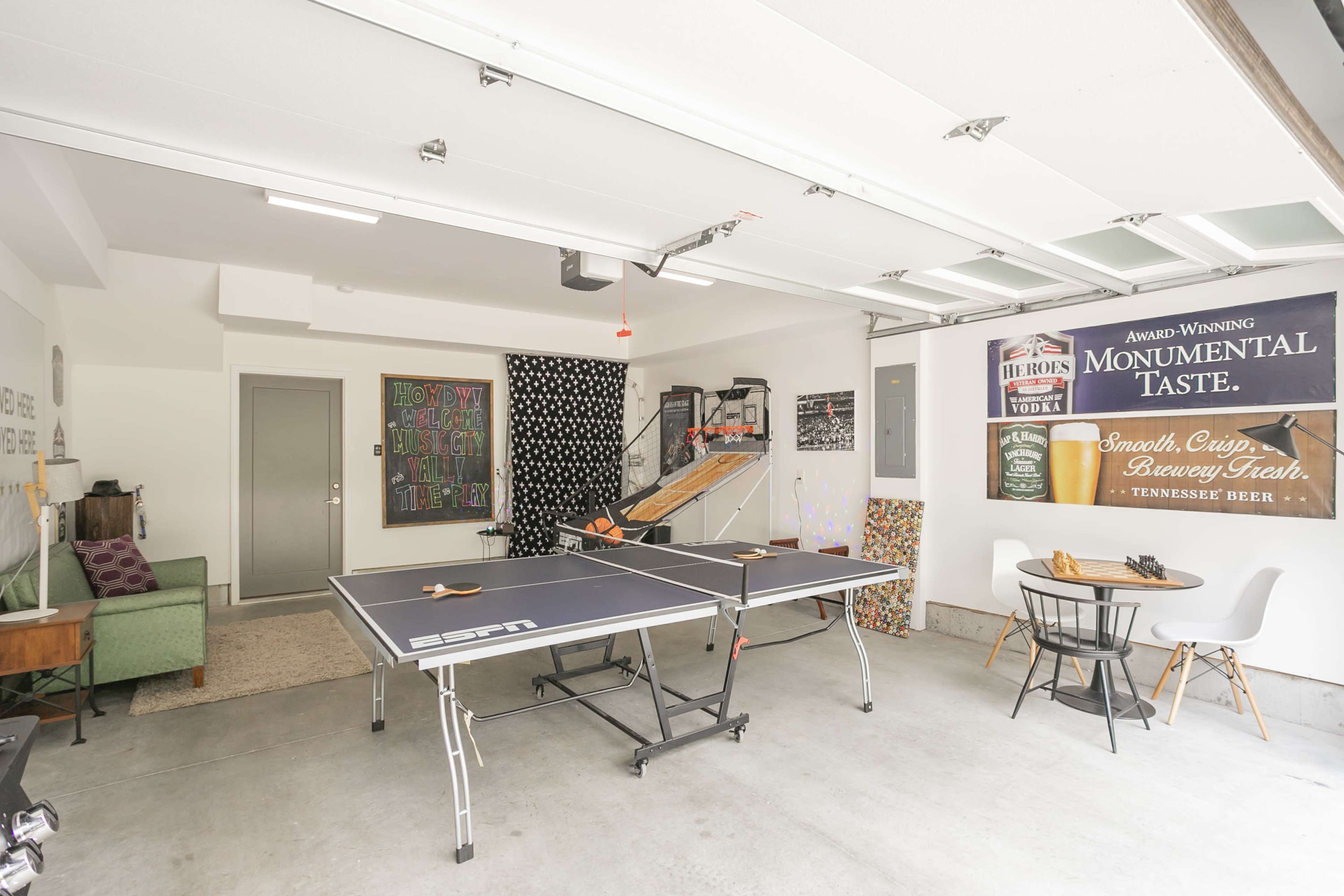 The image shows a garage with a ping pong table in the center, a lounge area with a green couch, and various decorative signs on the walls.