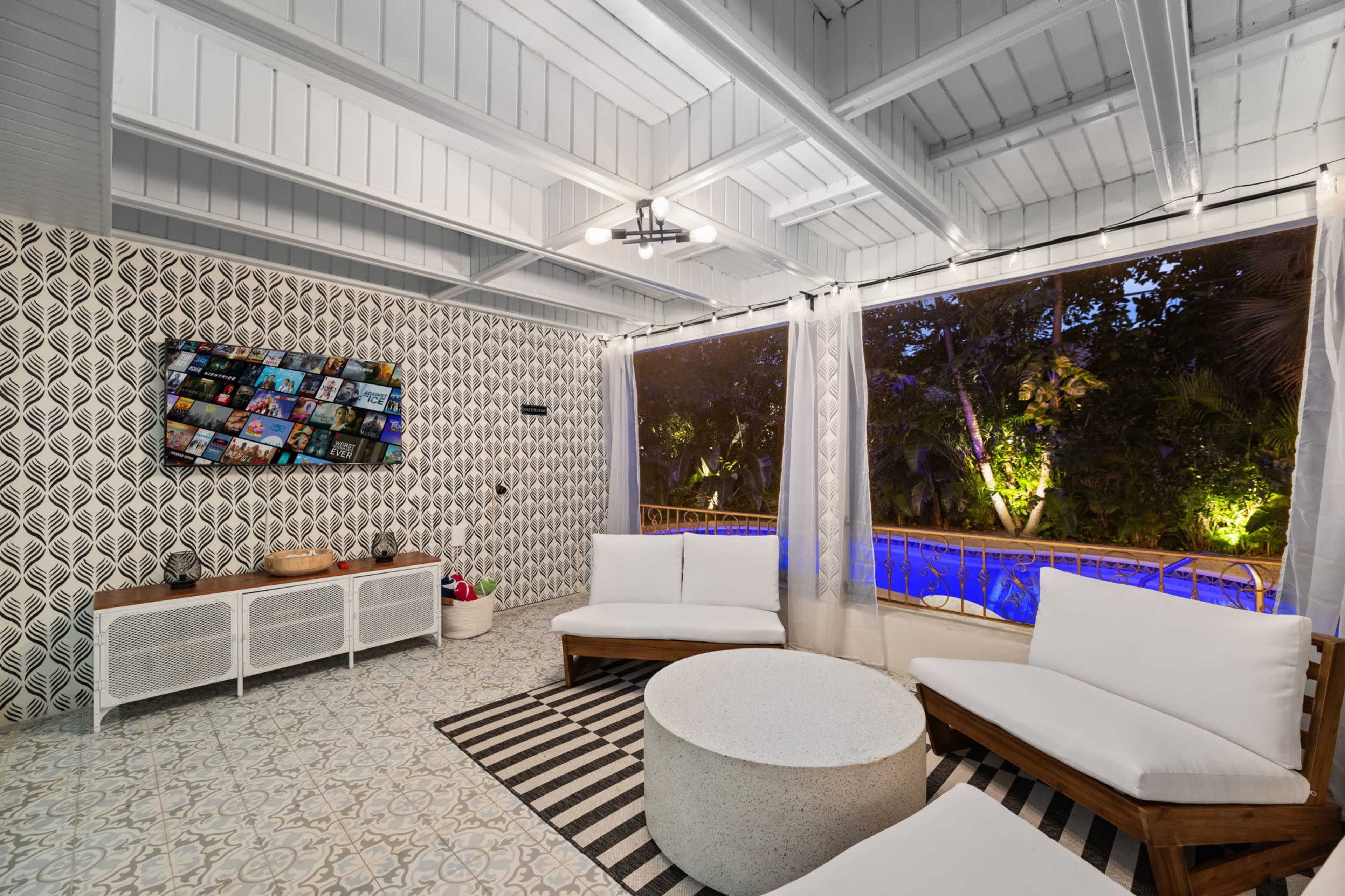 The image shows a modern living space with a seating area, a round coffee table, and a large window overlooking a lit outdoor area with a pool.