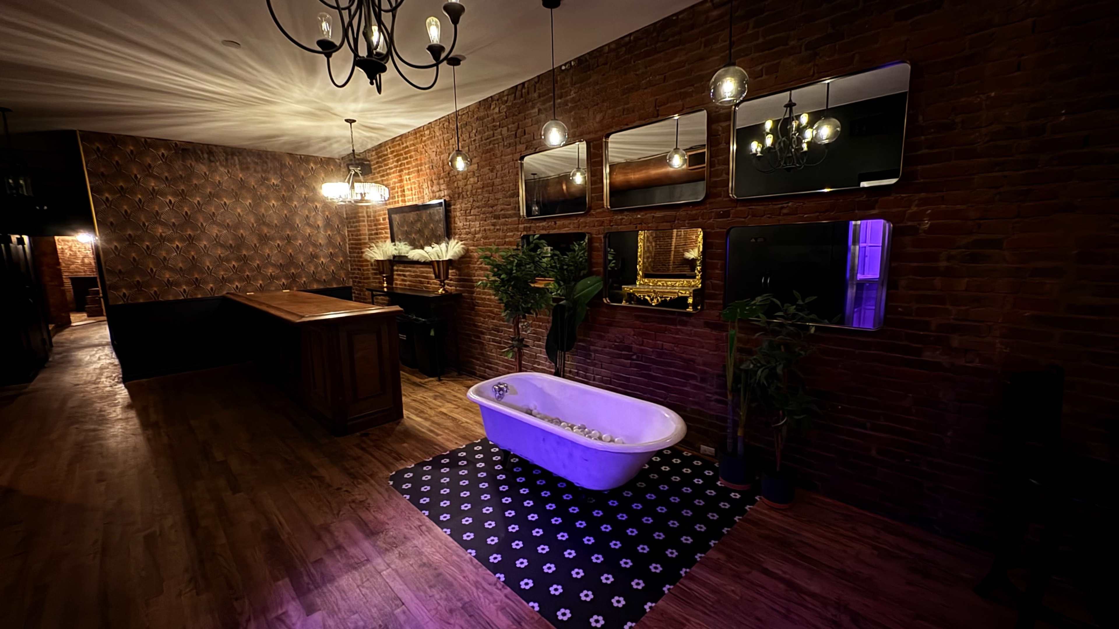 The image shows an interior space featuring a vintage bathtub, a bar with wooden cabinetry, decorative mirrors, plants, and a patterned rug against a backdrop of exposed brick walls and dim lighting.