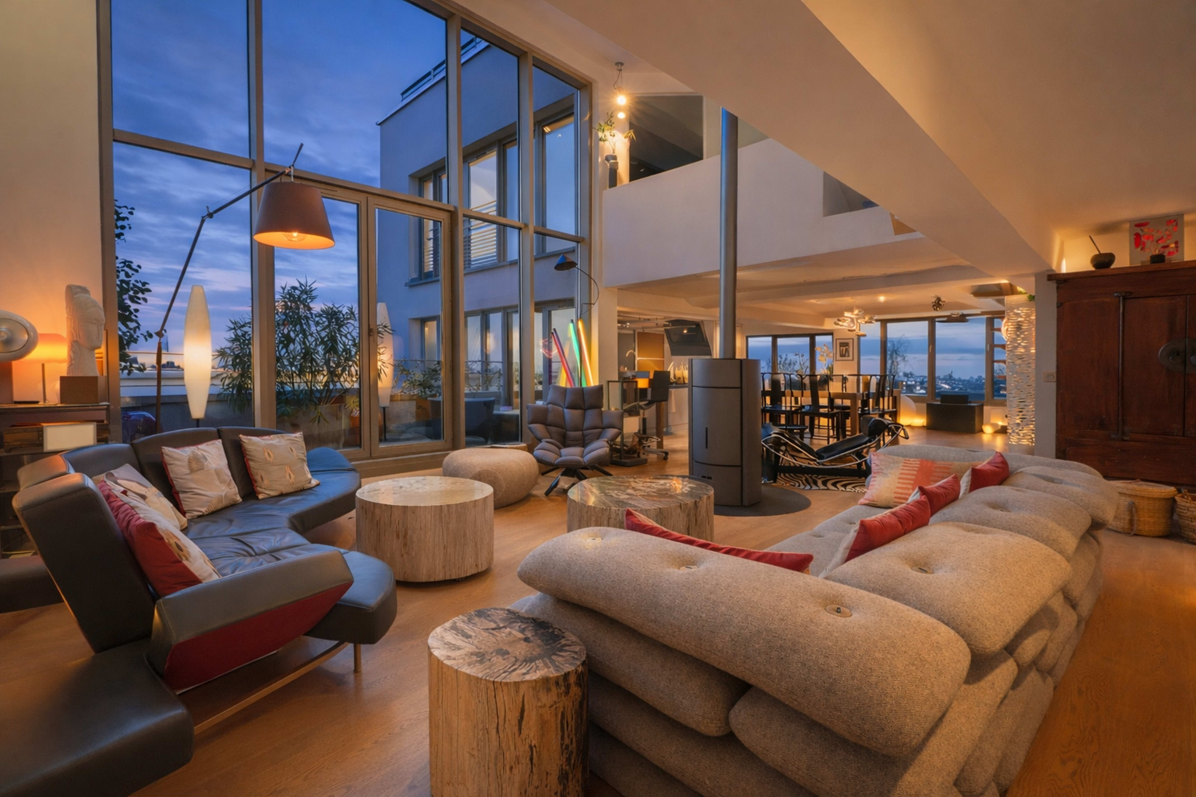 The image shows a modern living room with large windows, featuring contemporary furniture, wooden log tables, and an open layout that overlooks a scenic view.