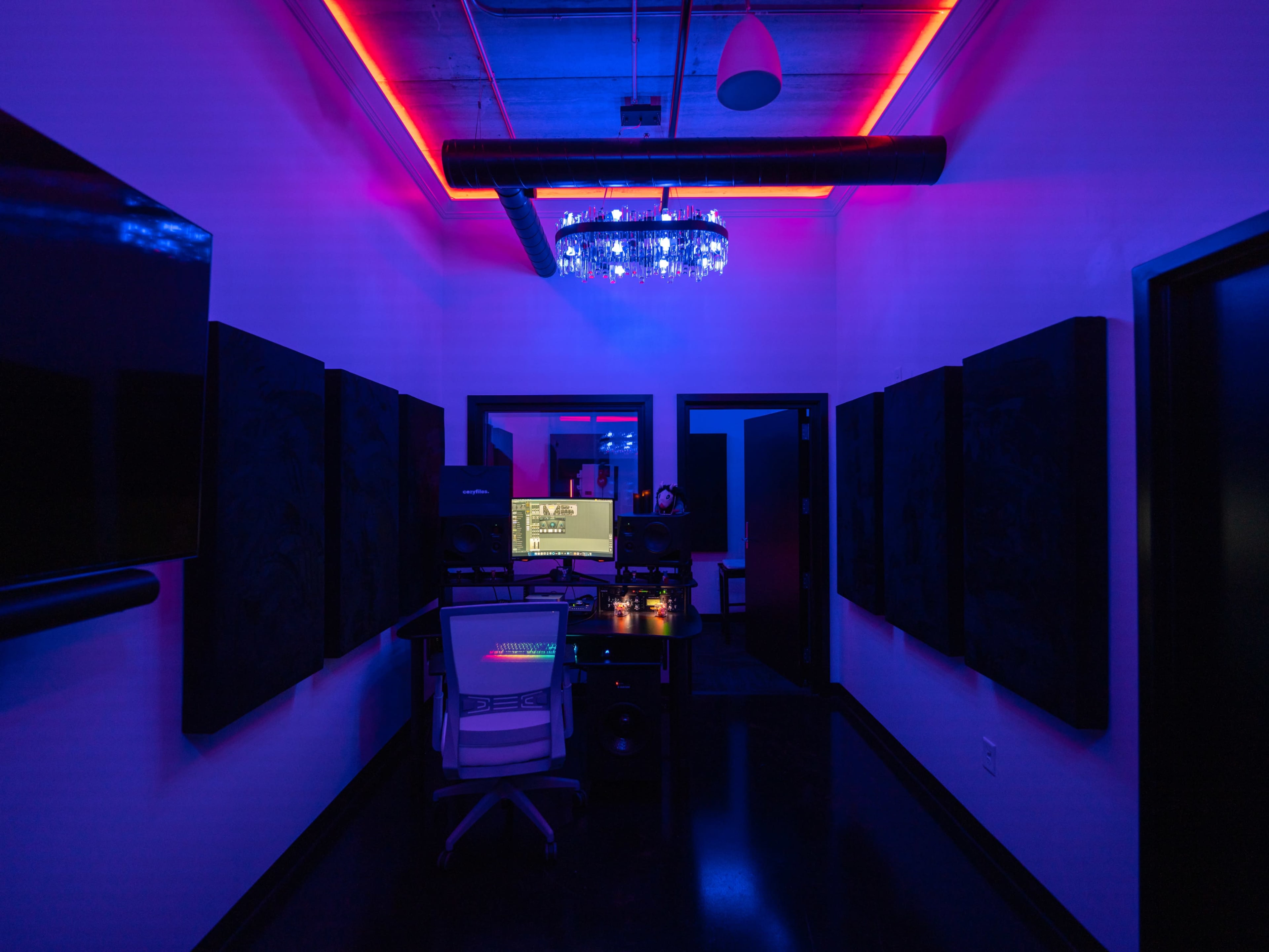 The image shows a dimly lit recording studio with neon lights, a computer setup on a desk, and soundproofing panels on the walls.