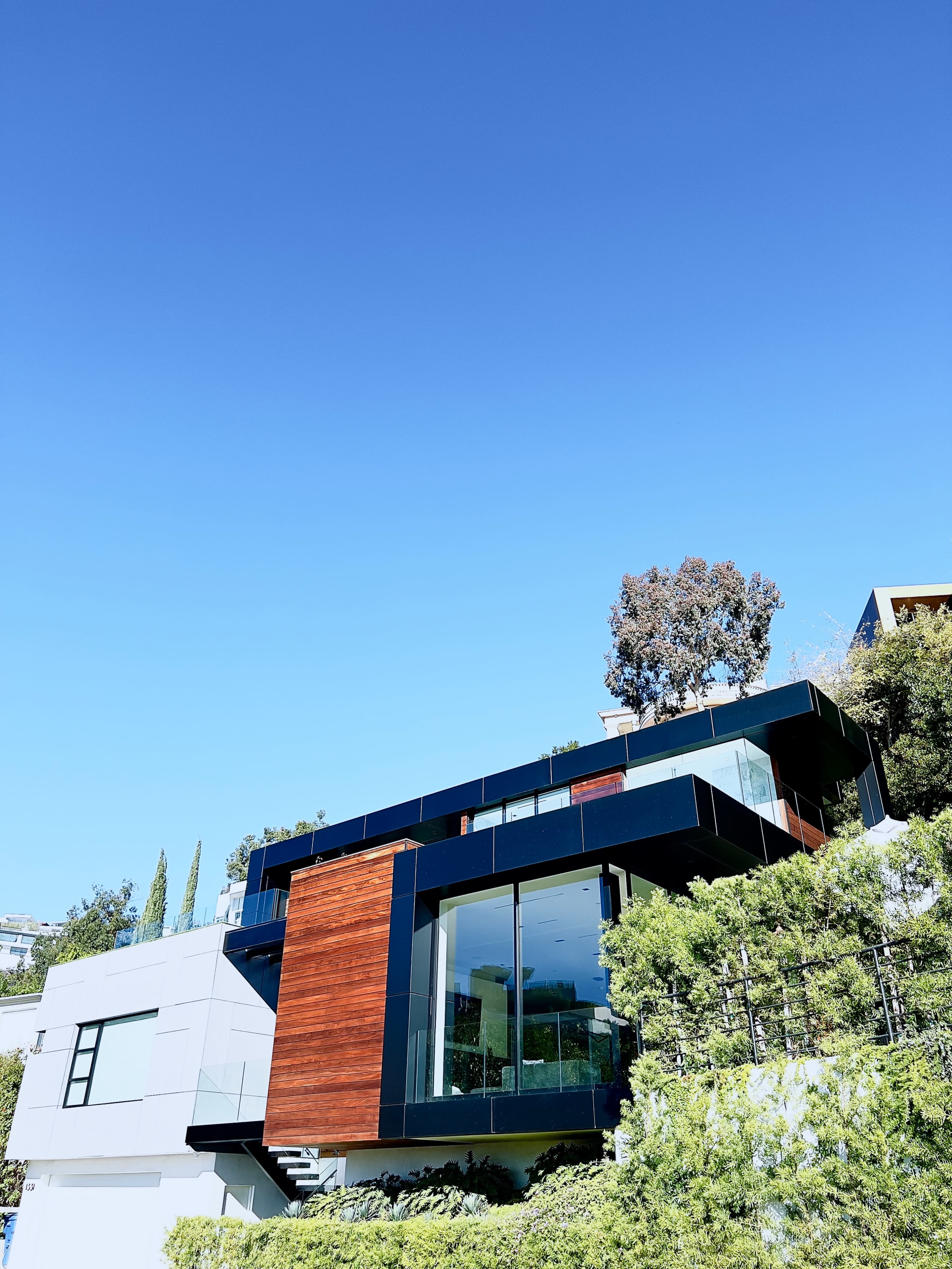 A contemporary house with large glass windows and a wooden facade, situated on a hillside under a clear blue sky.
