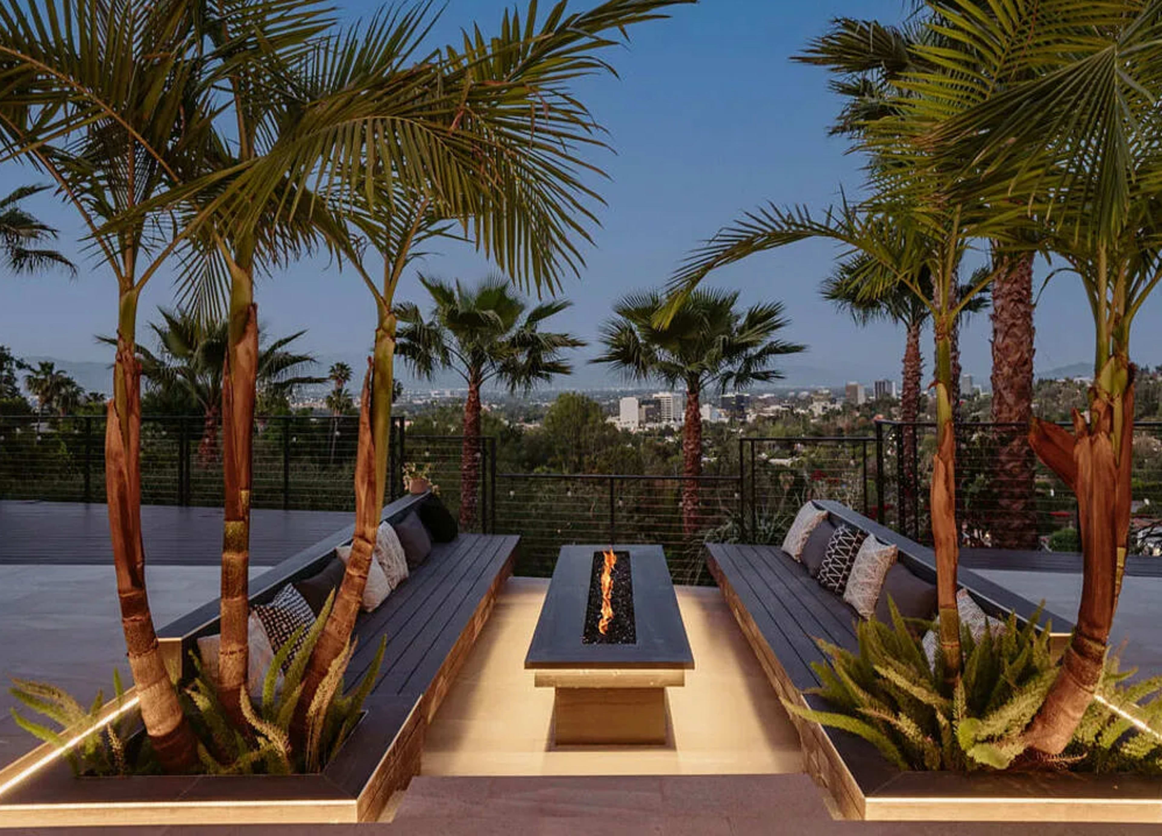 A modern outdoor seating area features a long fire pit surrounded by palm trees and lounge seating.