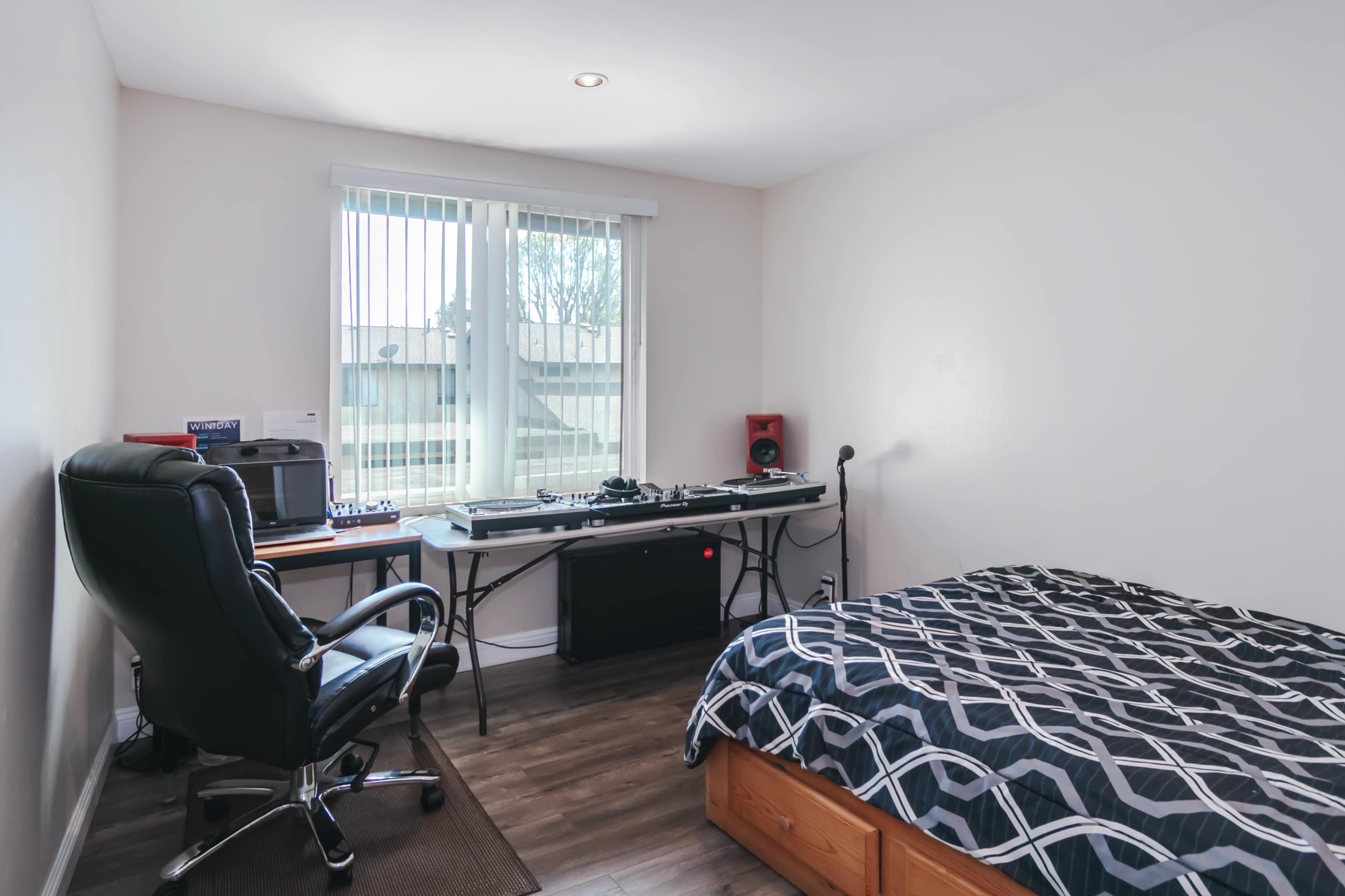 A simple bedroom with a bed, a desk and chair, and a DJ setup against the wall near a window.