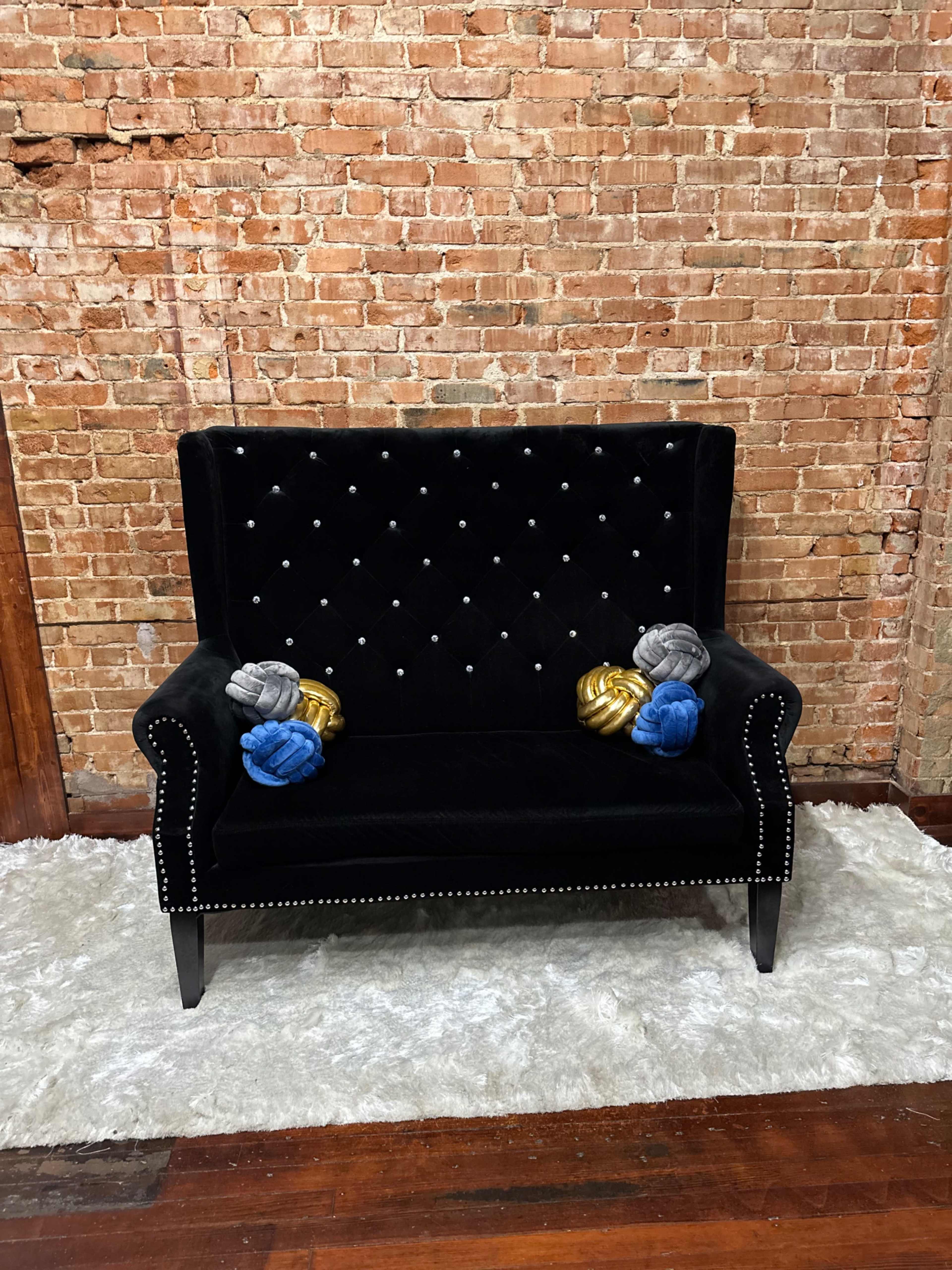A black tufted loveseat with decorative pillows sits on a white rug against a brick wall.