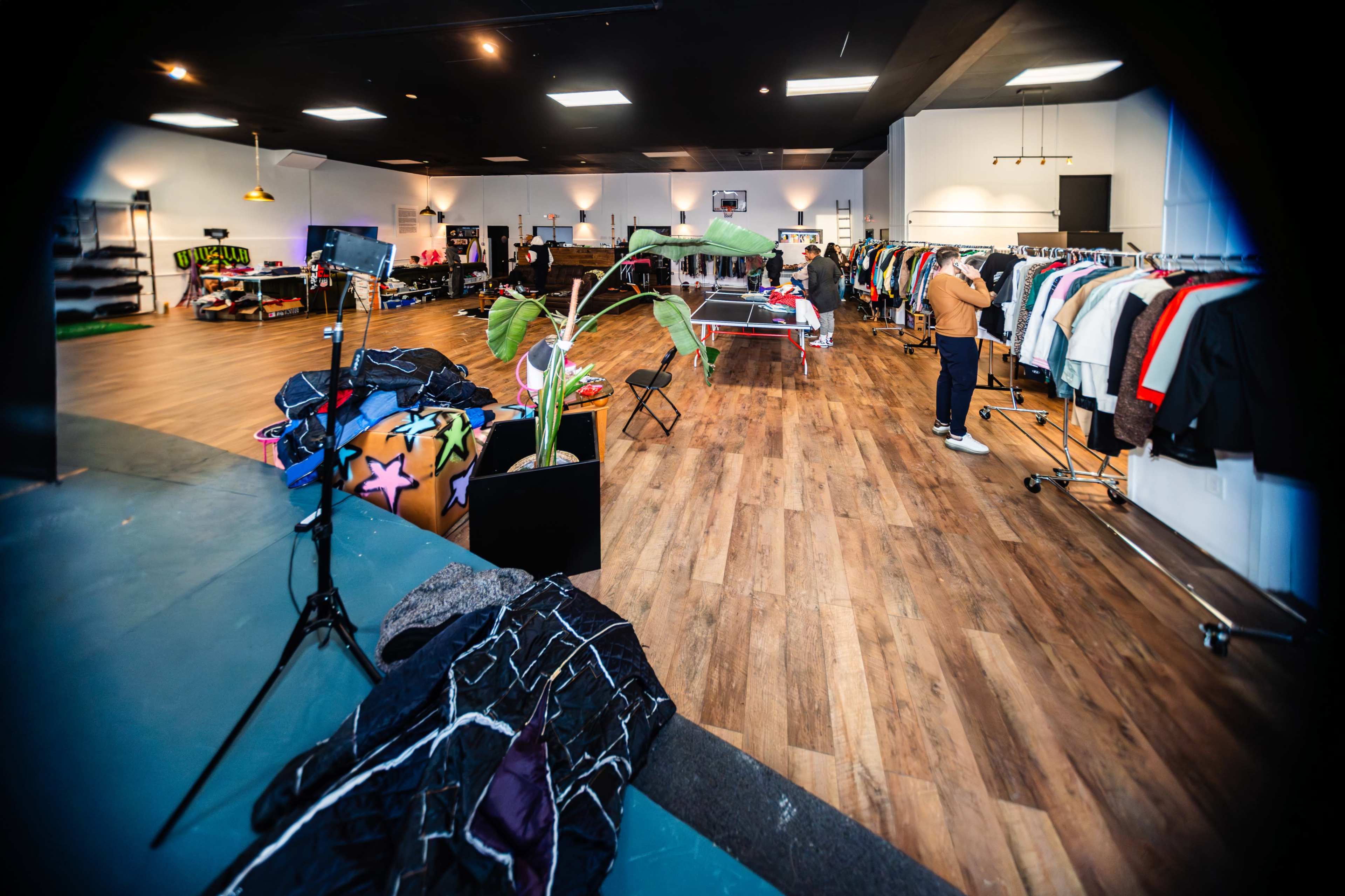 The image shows a clothing store interior with racks of garments, a seating area, and a green plant in the foreground.