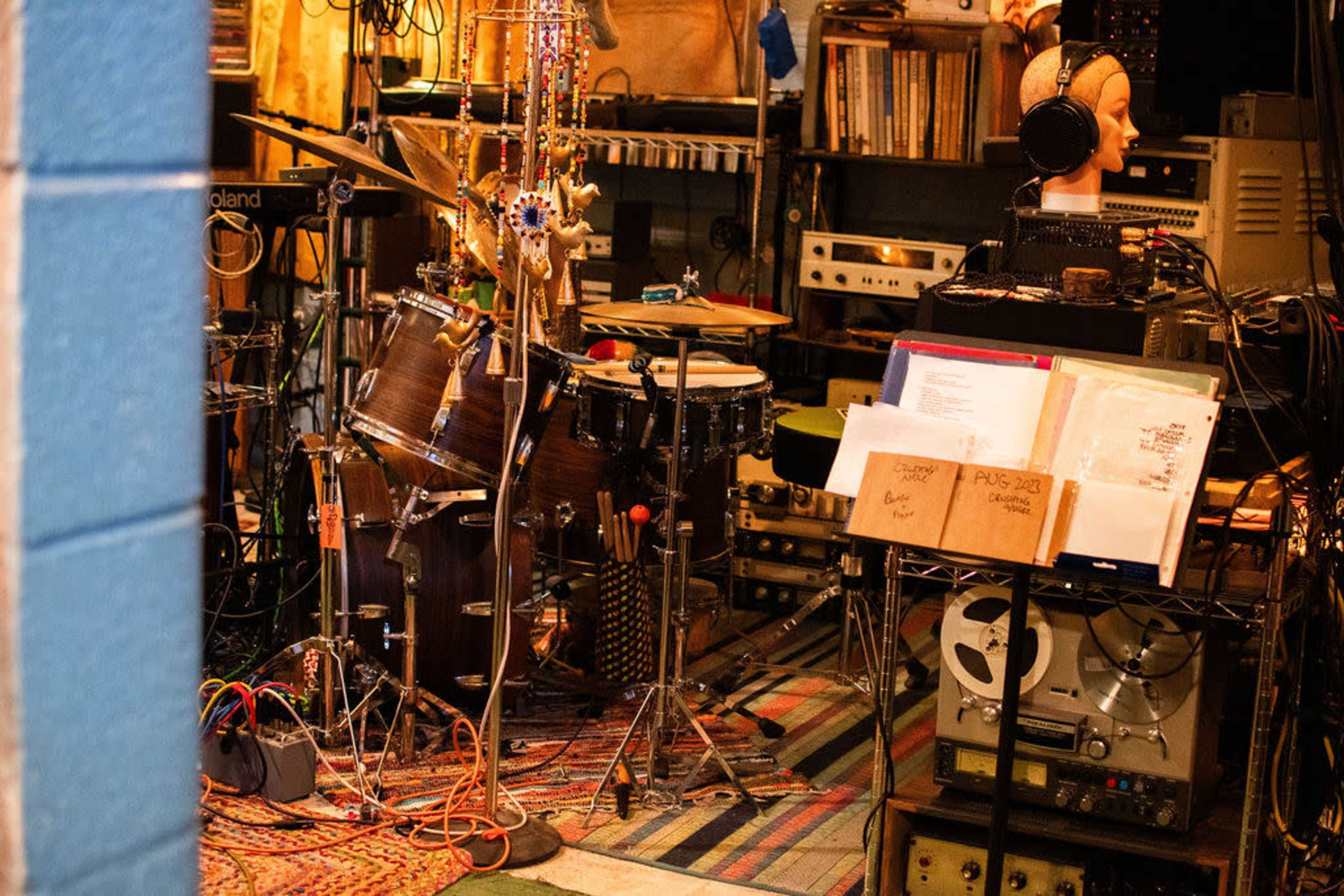 The image shows a cluttered music studio filled with various instruments, including a drum set and congas, alongside shelves of equipment and sheet music on a stand.
