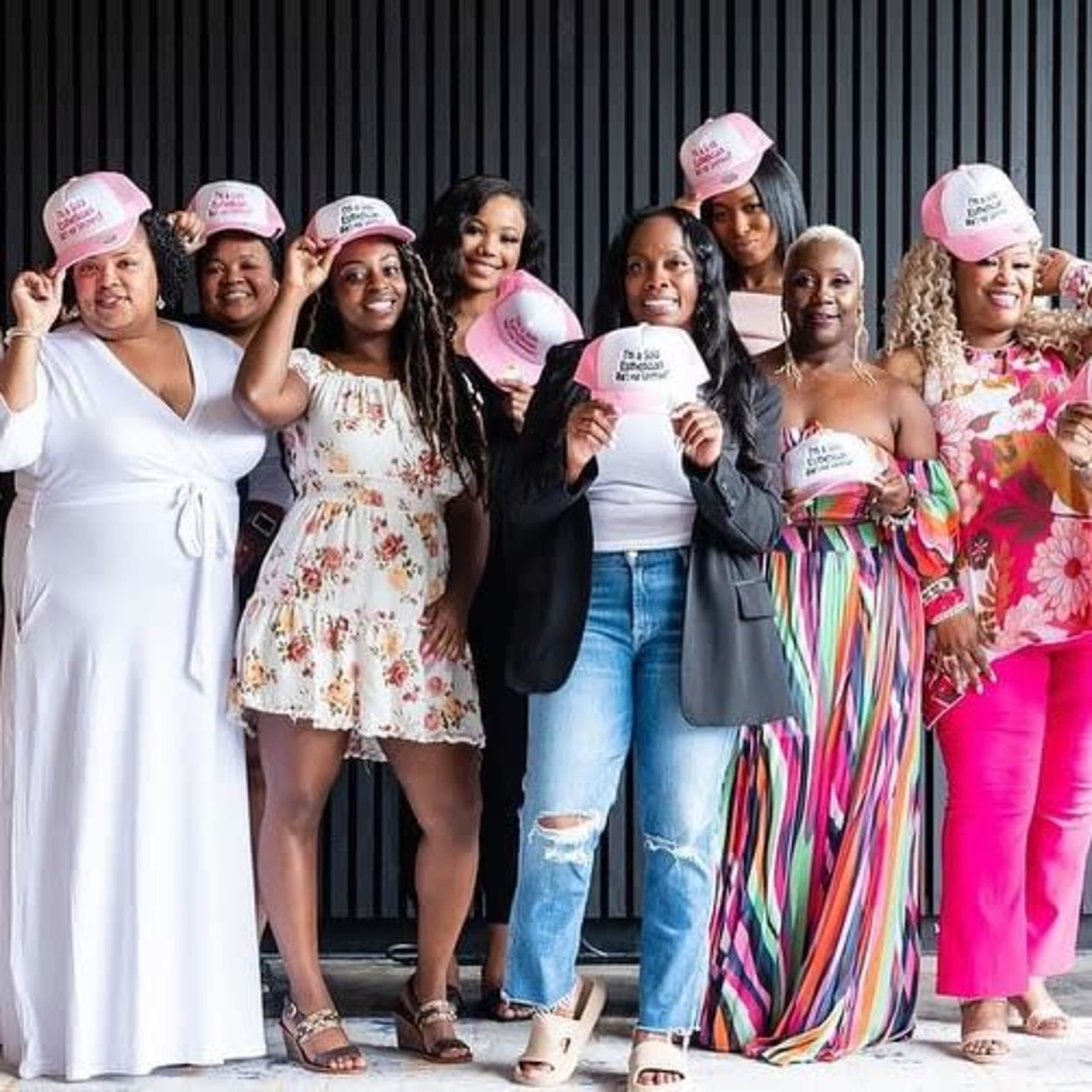 A group of eight women are posing together, wearing pink hats and showcasing a variety of colorful outfits.