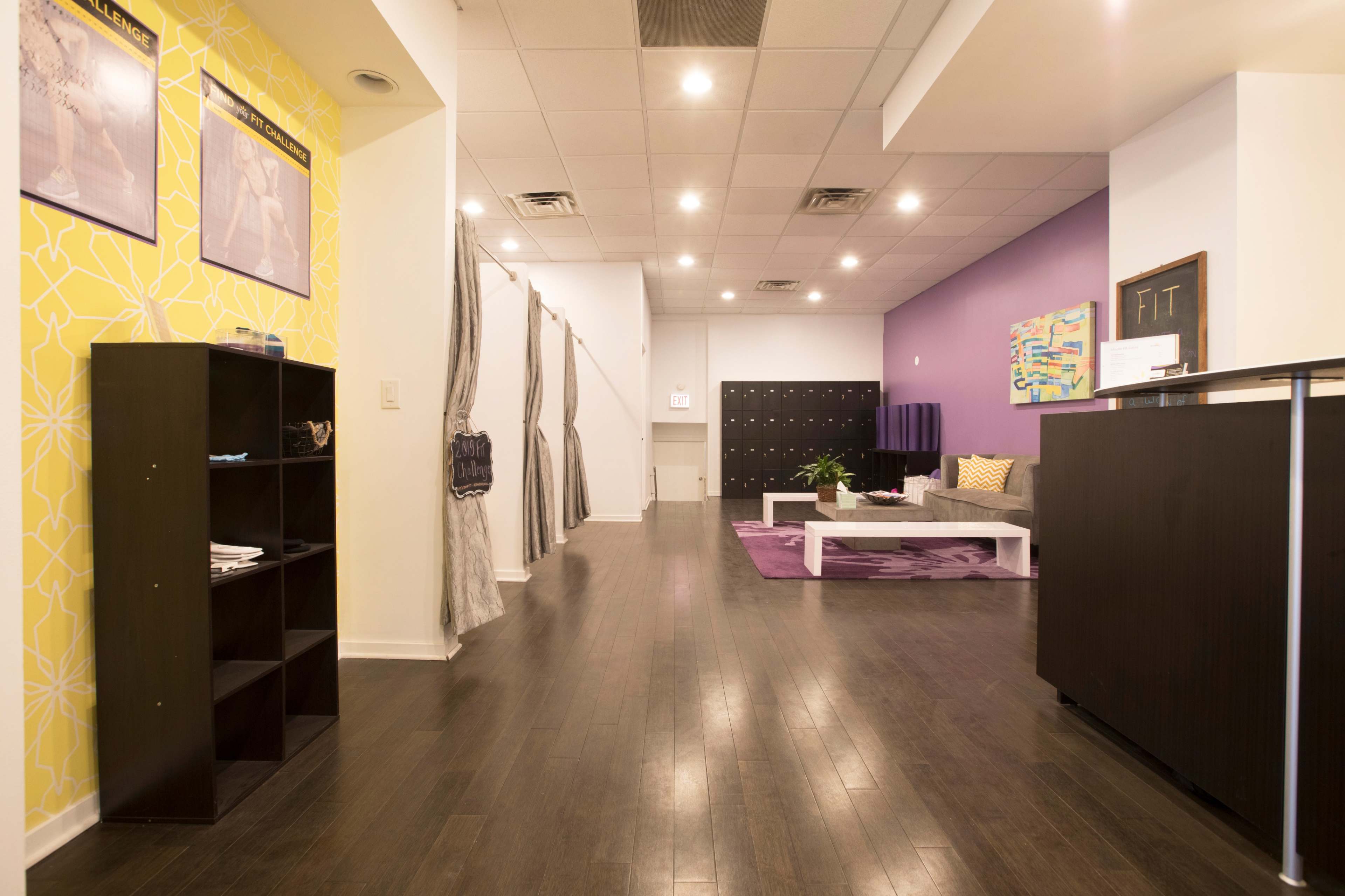 The image shows a spacious interior of a modern reception area with a bright yellow accent wall, dark wooden flooring, and a seating area featuring a purple rug and decorative cushions.