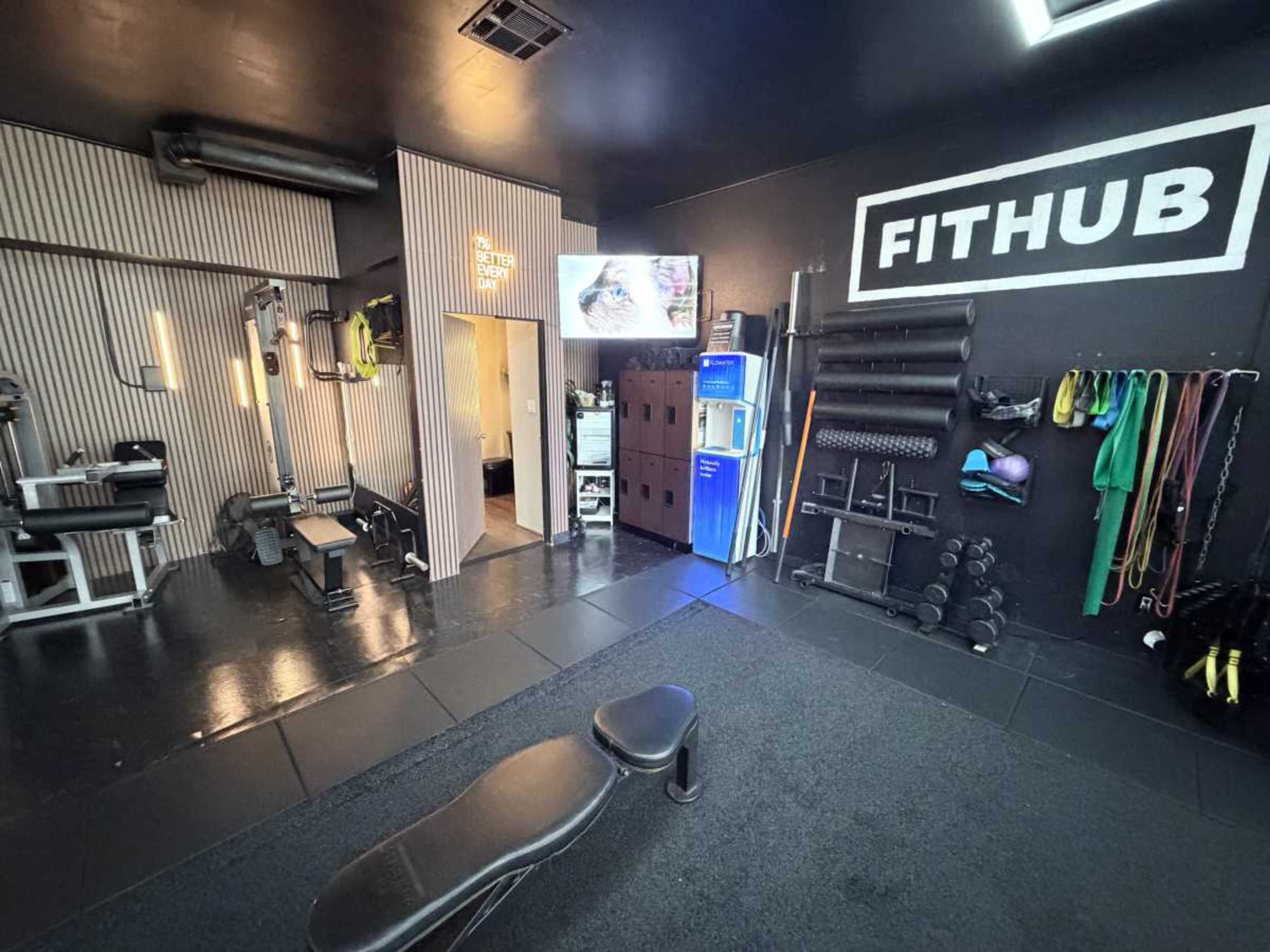 The image shows a compact gym space featuring exercise equipment, a television, storage lockers, and fitness accessories against a black and striped wall.