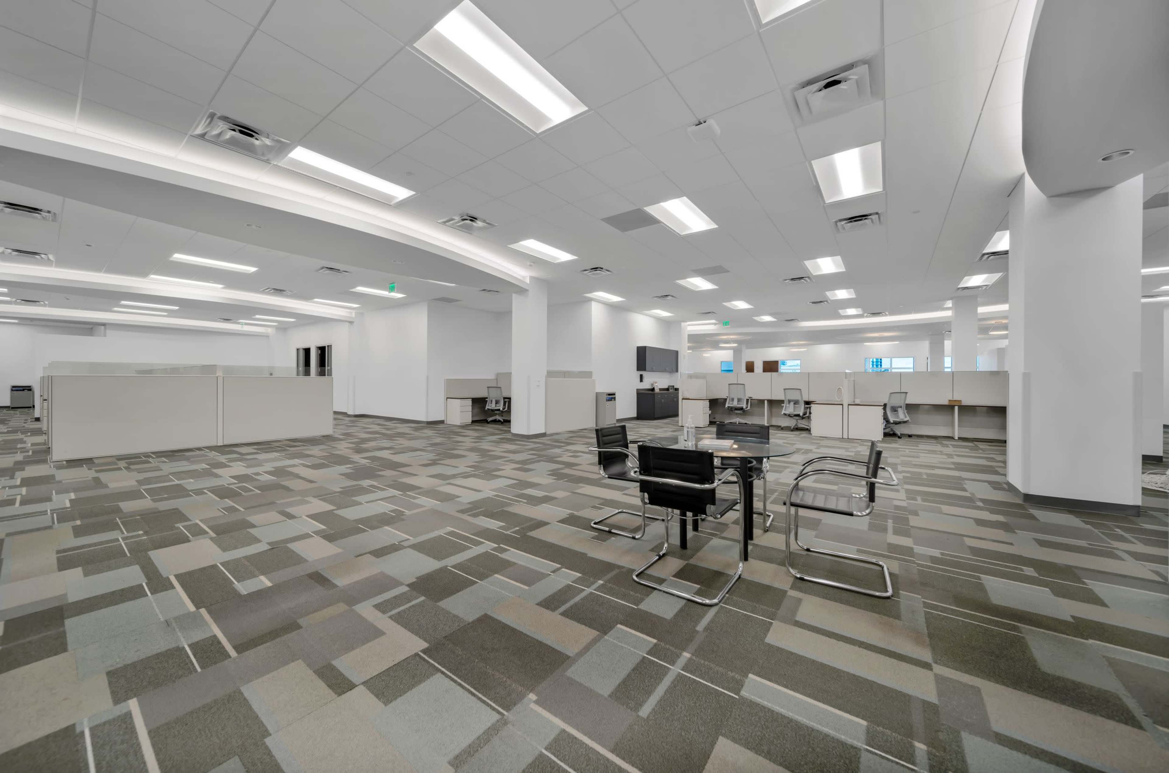 An empty office space featuring cubicles and a seating area with a table and chairs on a tiled floor.