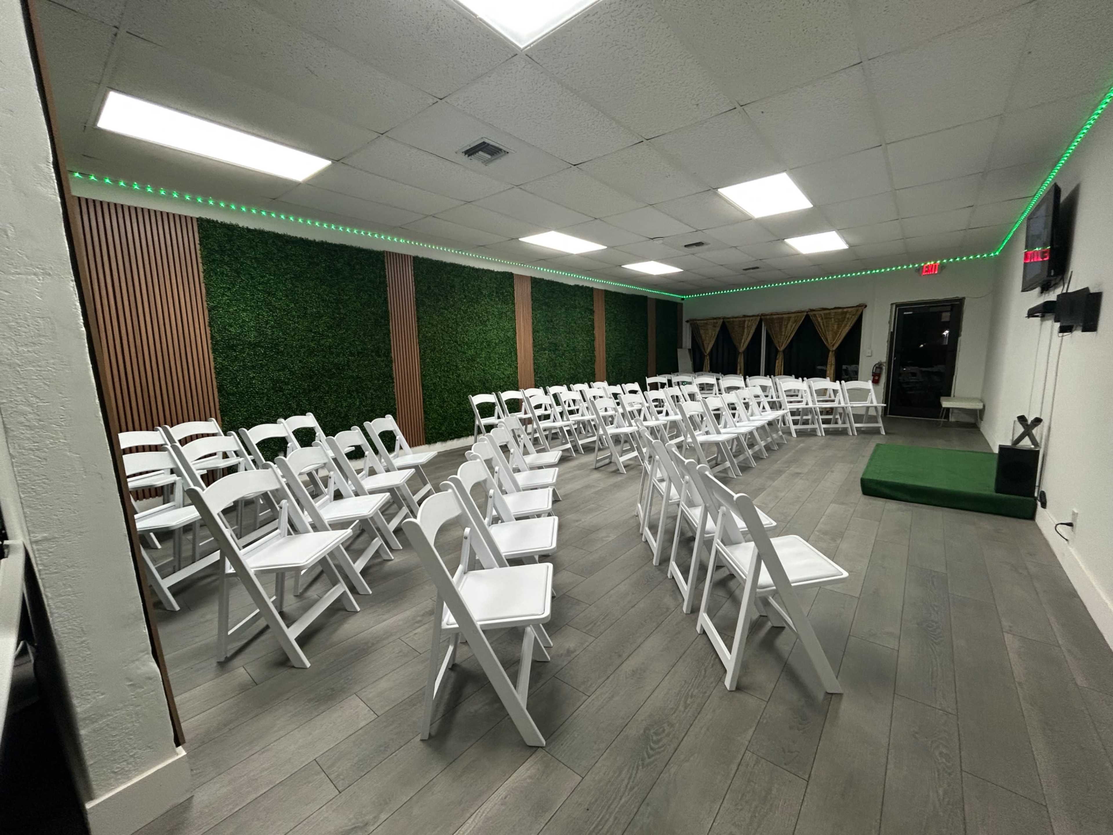 The image shows a room set up with white folding chairs arranged in rows facing a small green stage area, with greenery-covered walls and ambient lighting.