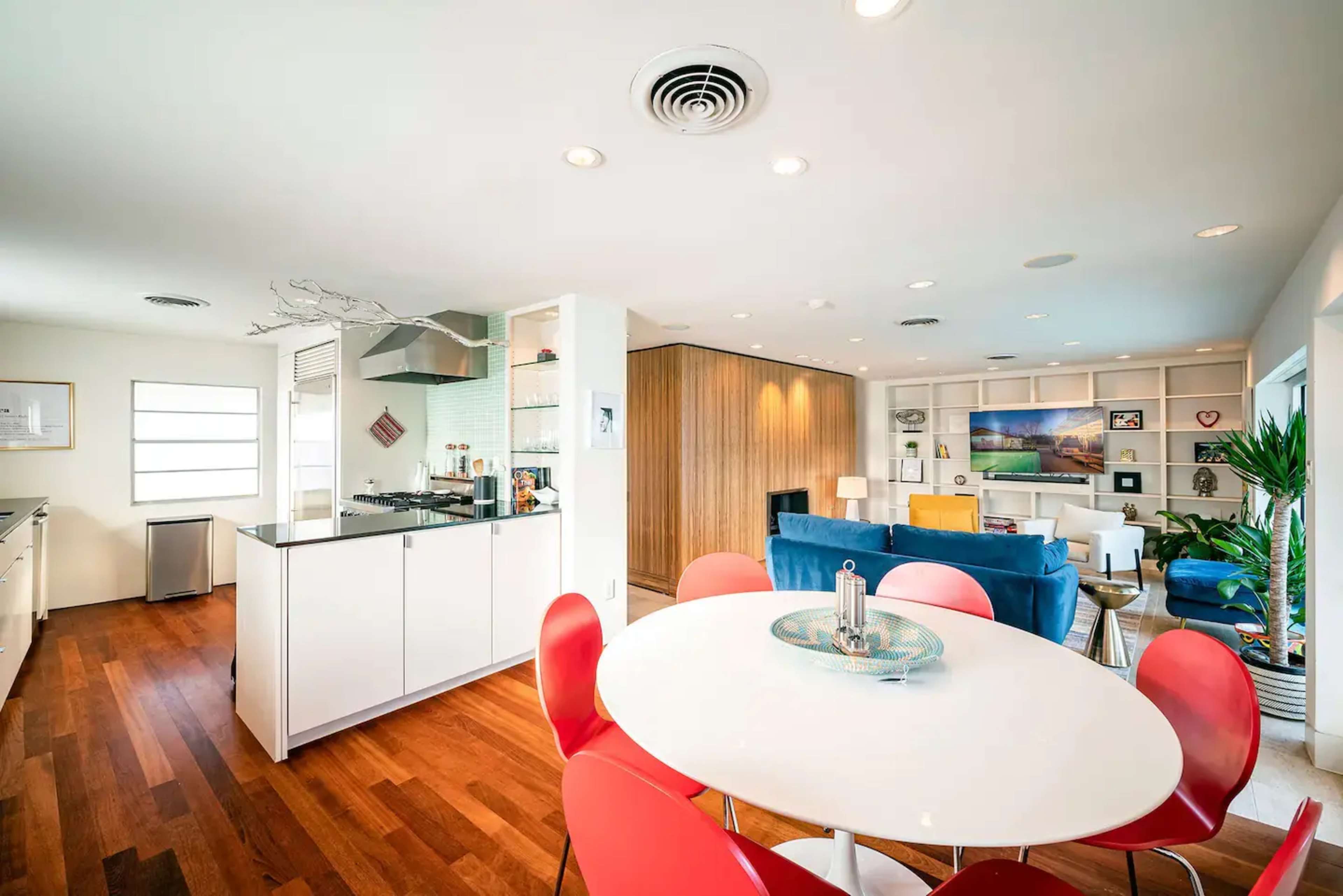 A modern kitchen and living area feature an open layout with a round dining table and red chairs adjacent to a blue sofa and a wooden accent wall.