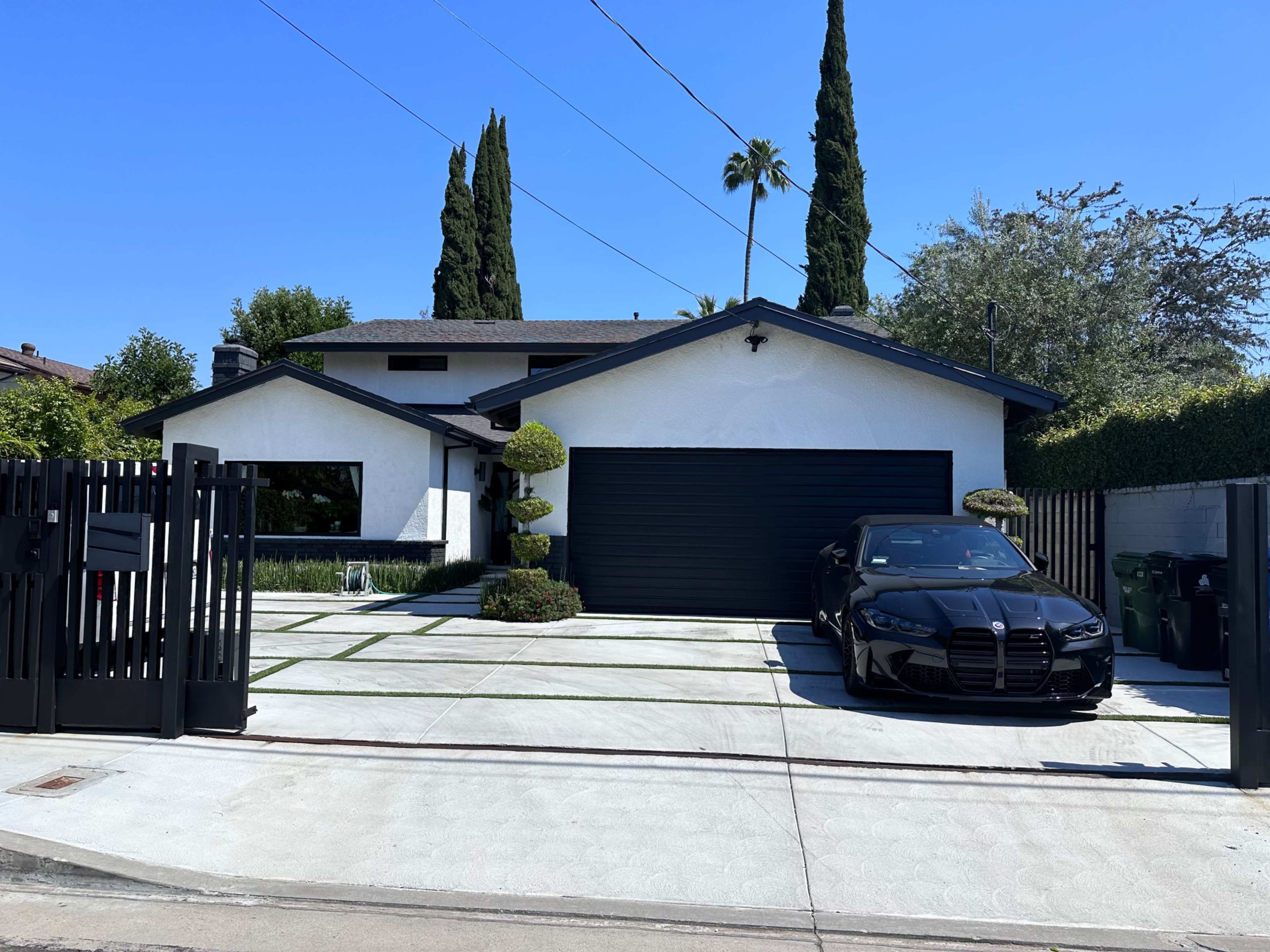A modern white house with a black garage door and a black car parked in front, framed by neatly trimmed shrubs and palm trees.