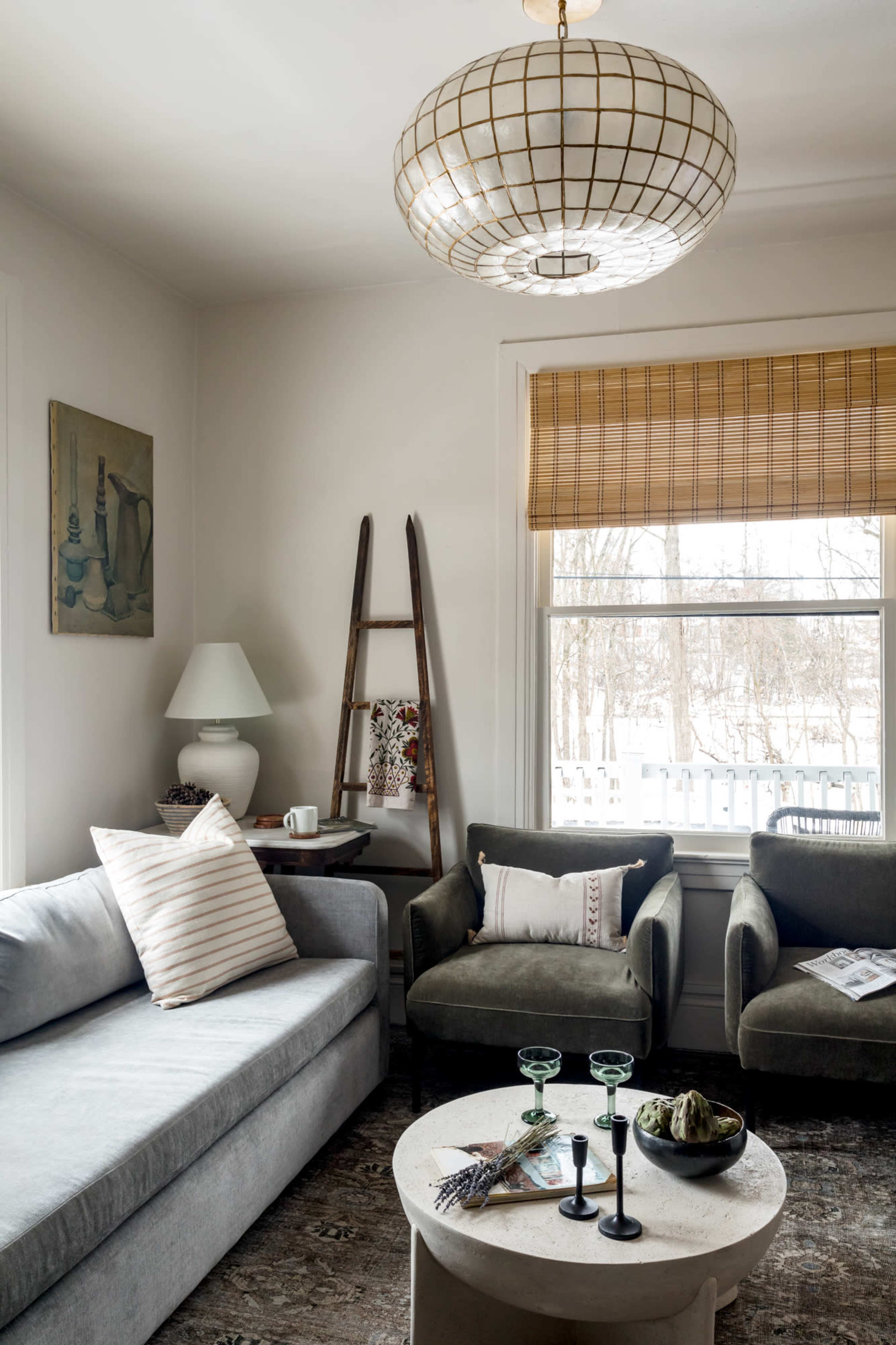 A cozy living room features a gray sofa, two green chairs, a round coffee table, and a large pendant light, with a window draped in bamboo shades.
