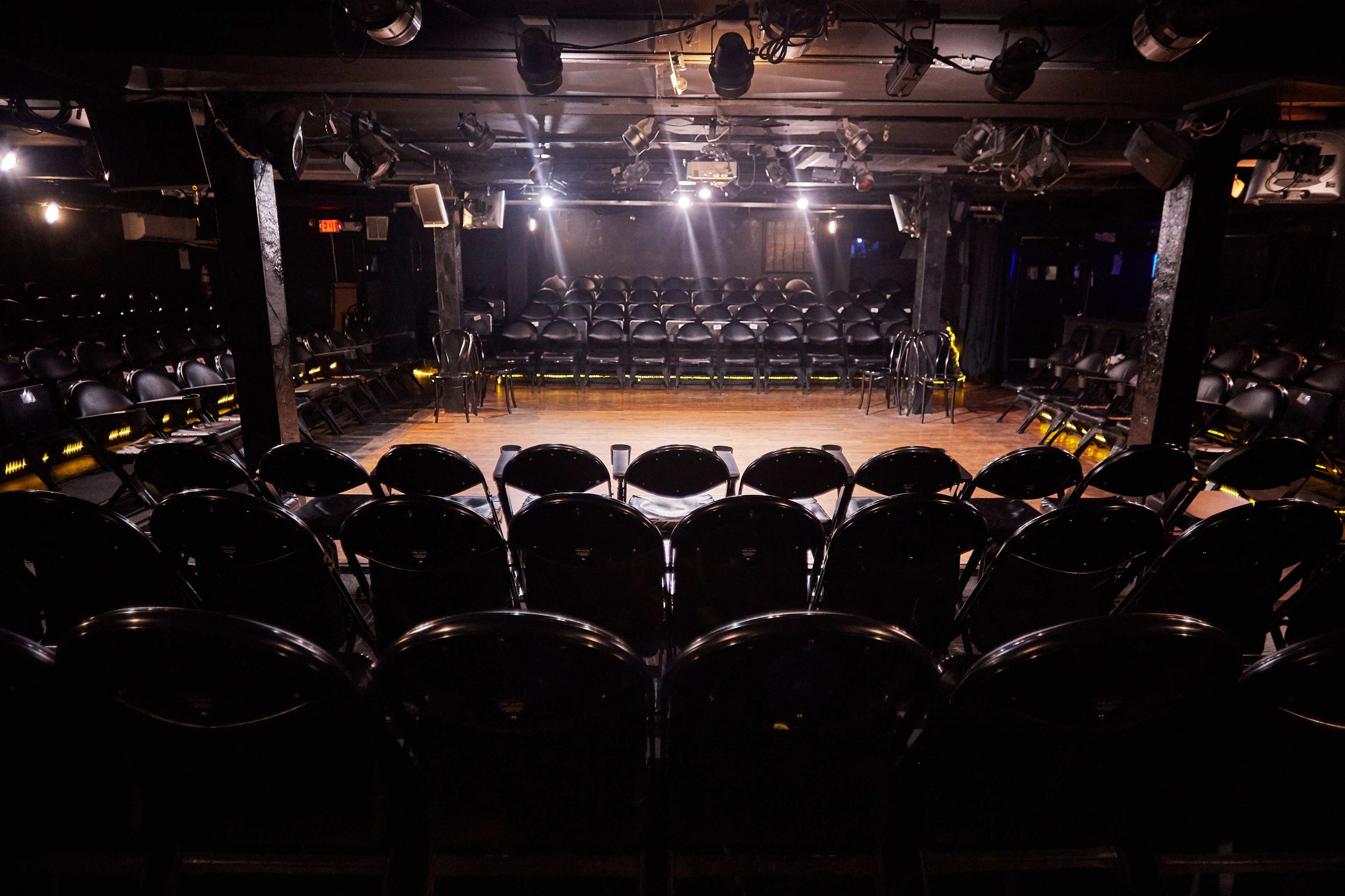 A dimly lit comedy club features a wooden stage surrounded by rows of black chairs.