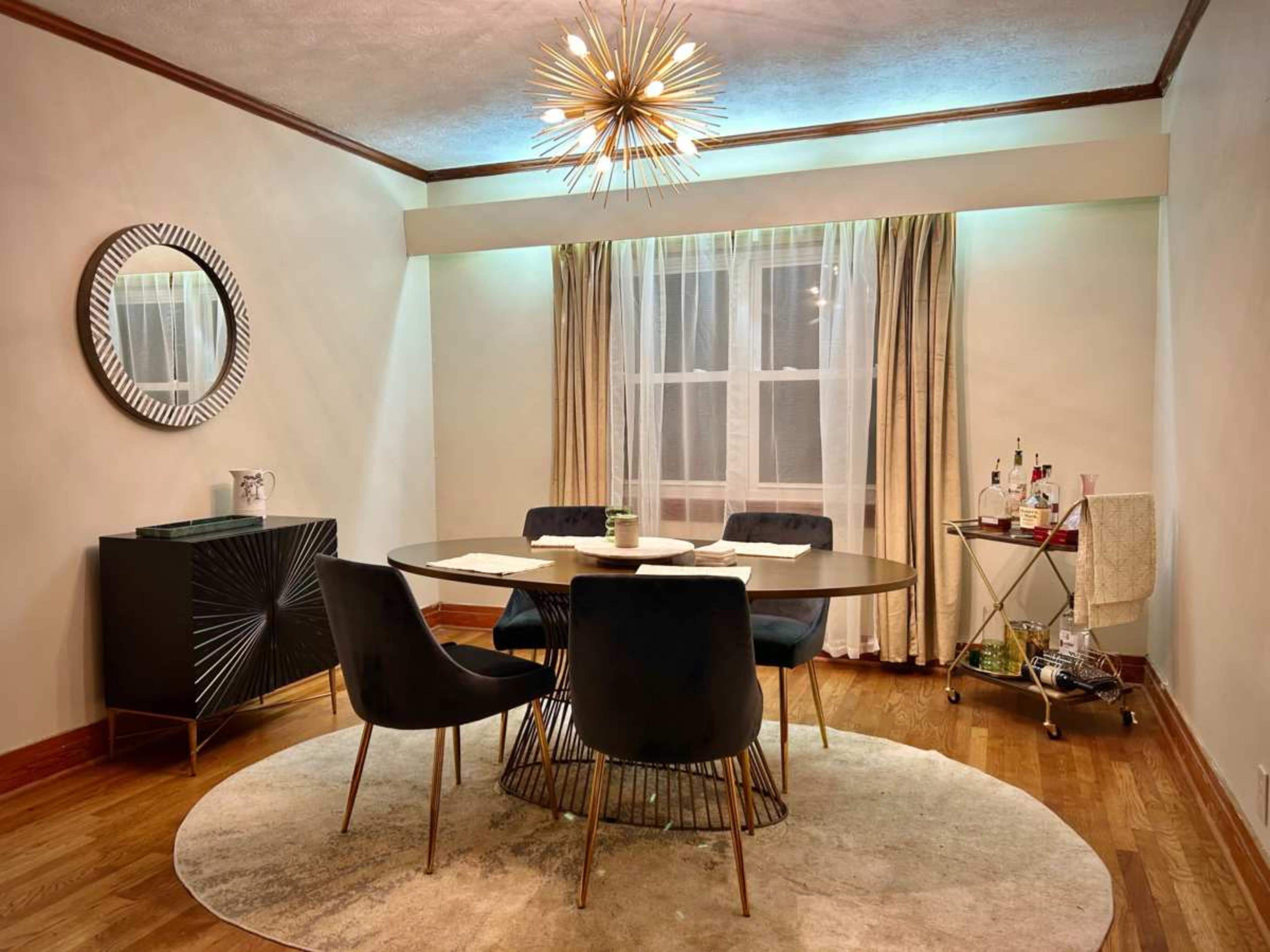 The image shows a dining room featuring a round table with four chairs, a mirrored wall decoration, and a bar cart in a corner.