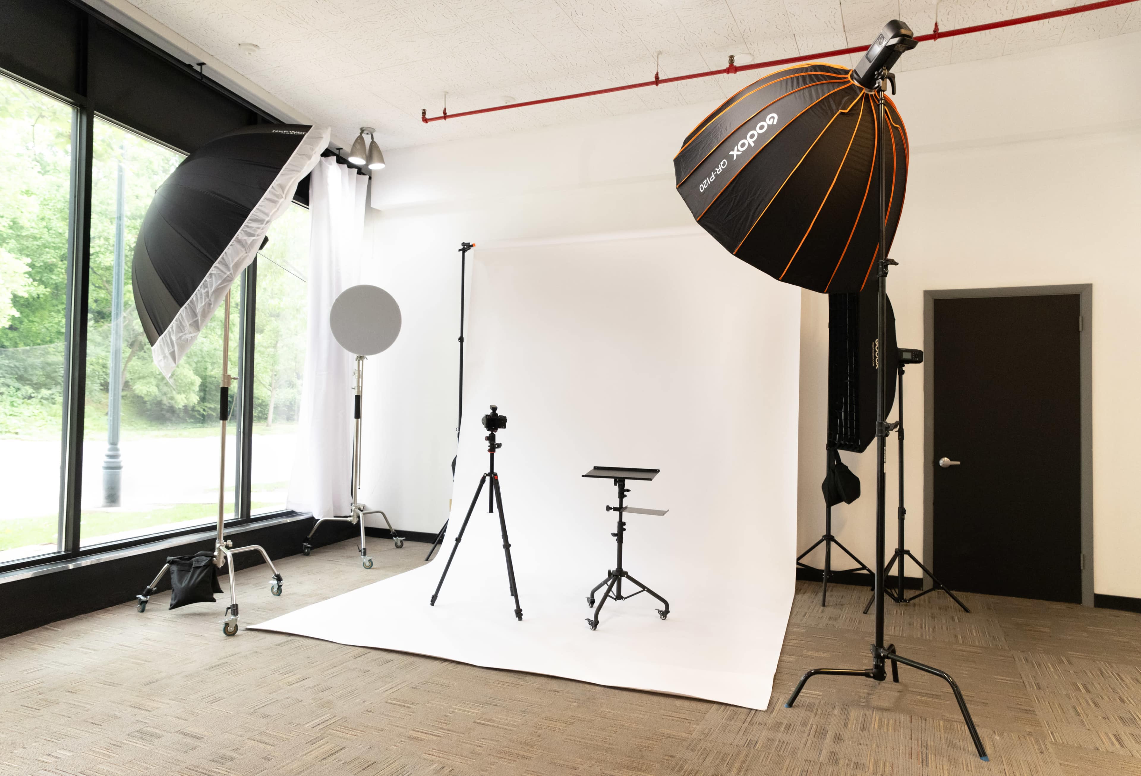 The image shows a photography studio setup featuring a white backdrop, a tripod with a camera, and various lighting equipment.