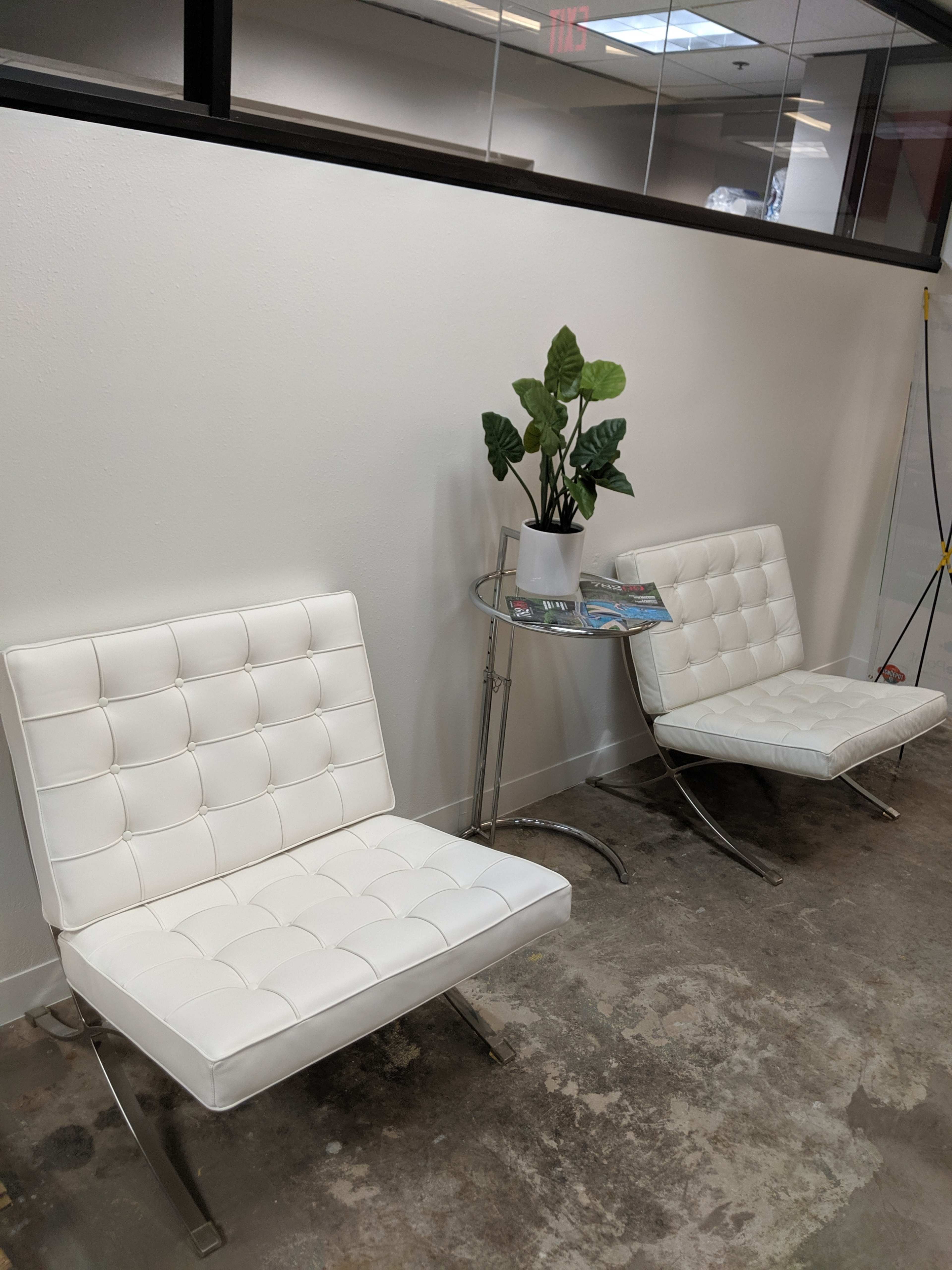 Two white chairs with tufted seats placed beside a small round table featuring a potted plant and a magazine, against a white wall in an office setting.