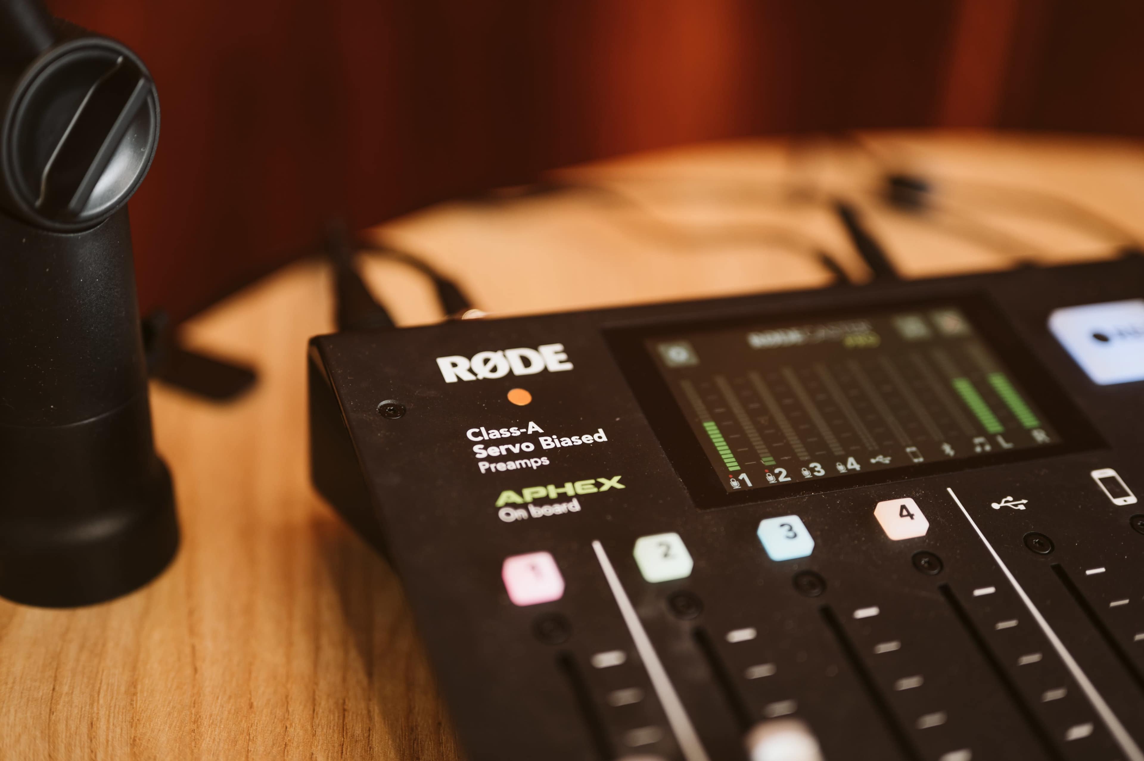 The image shows a close-up of a Rode audio mixing console featuring various controls and a display screen on a wooden surface.