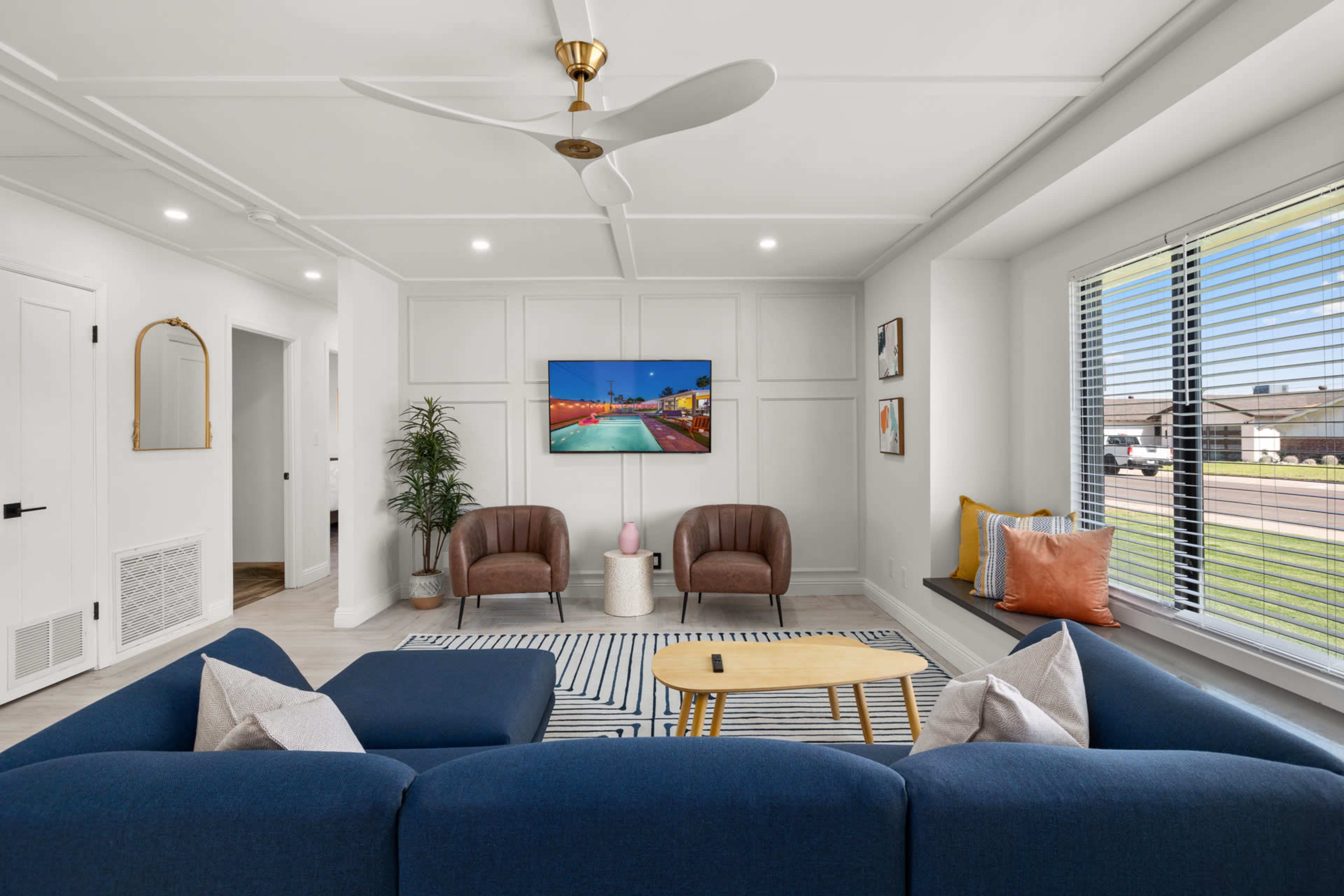 A modern living room features a blue sectional sofa, two brown chairs, a round coffee table, and a television mounted on the wall.
