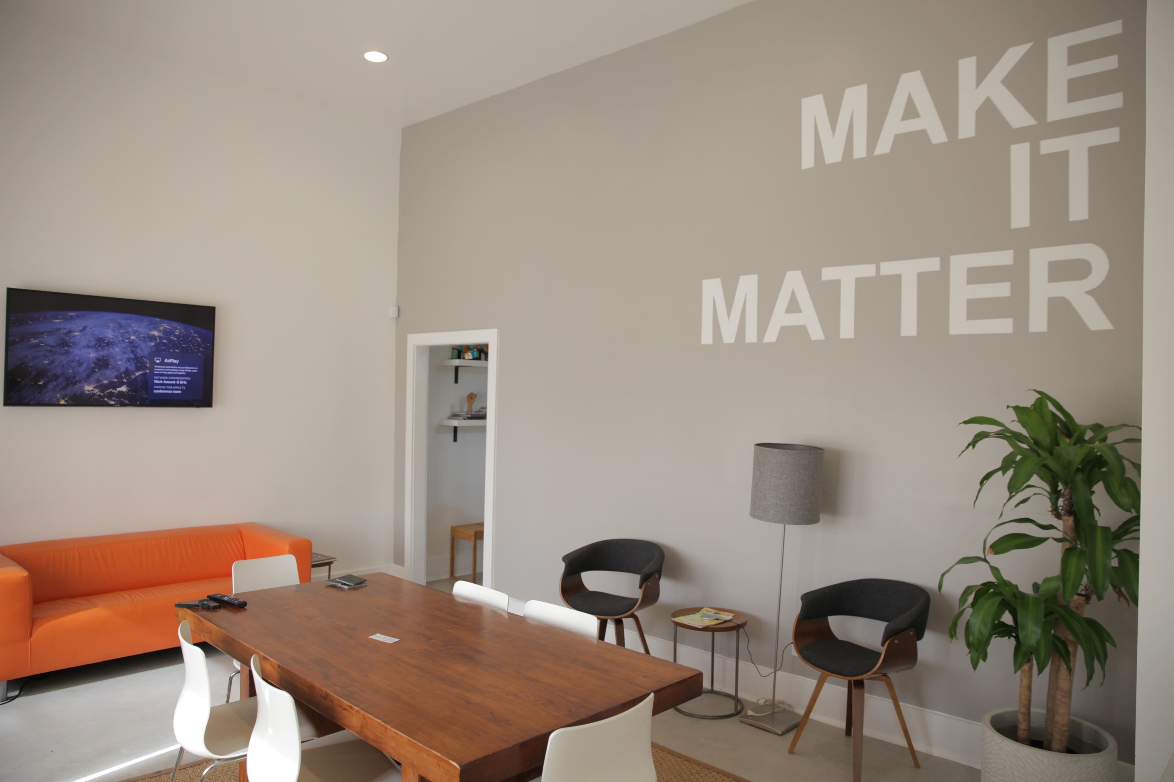 A modern lounge area features an orange sofa, a wooden table surrounded by chairs, and a wall with the phrase "MAKE IT MATTER" displayed prominently.