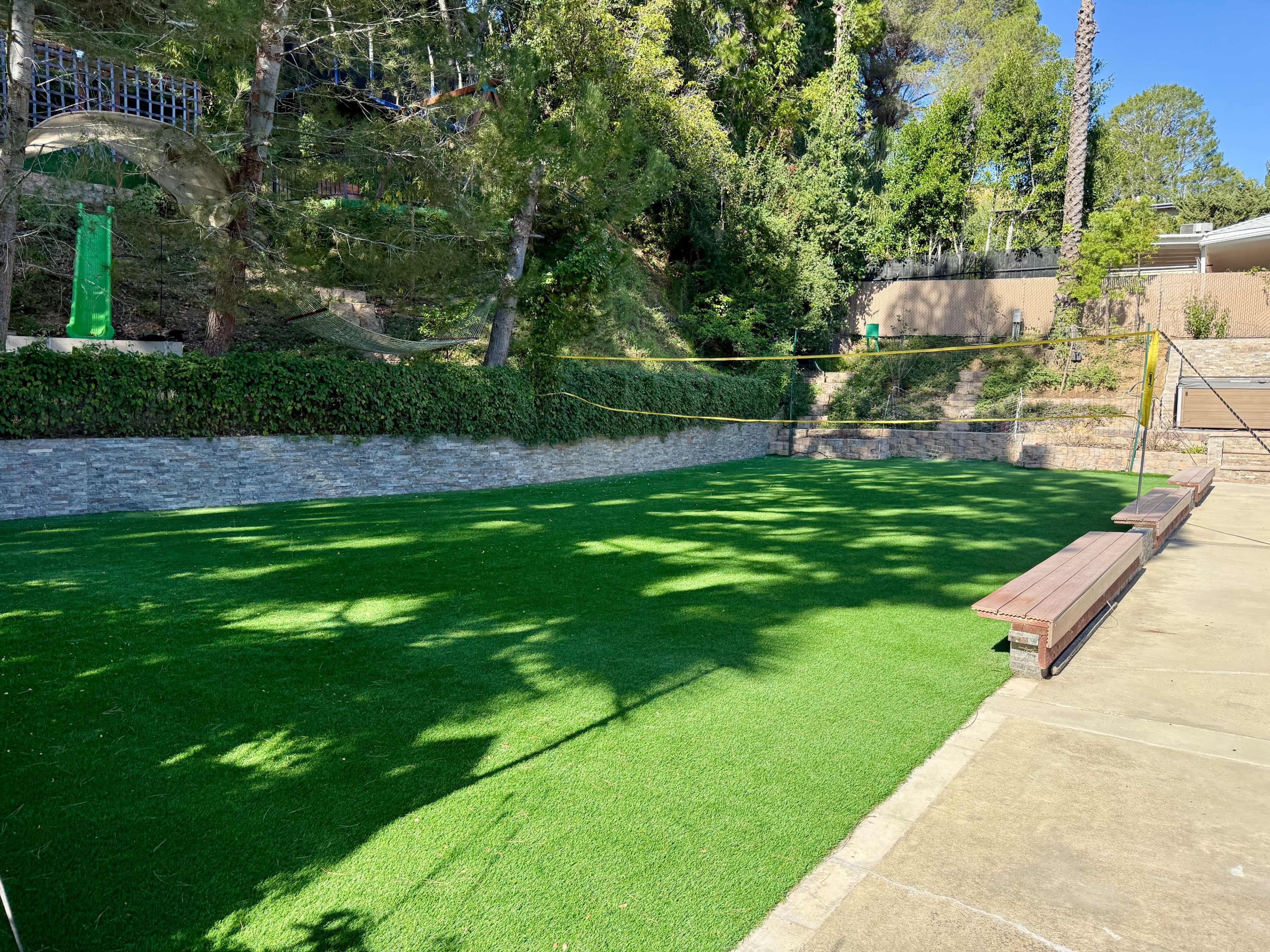 The image shows a grassy area with artificial turf, surrounded by trees and a concrete path with benches.