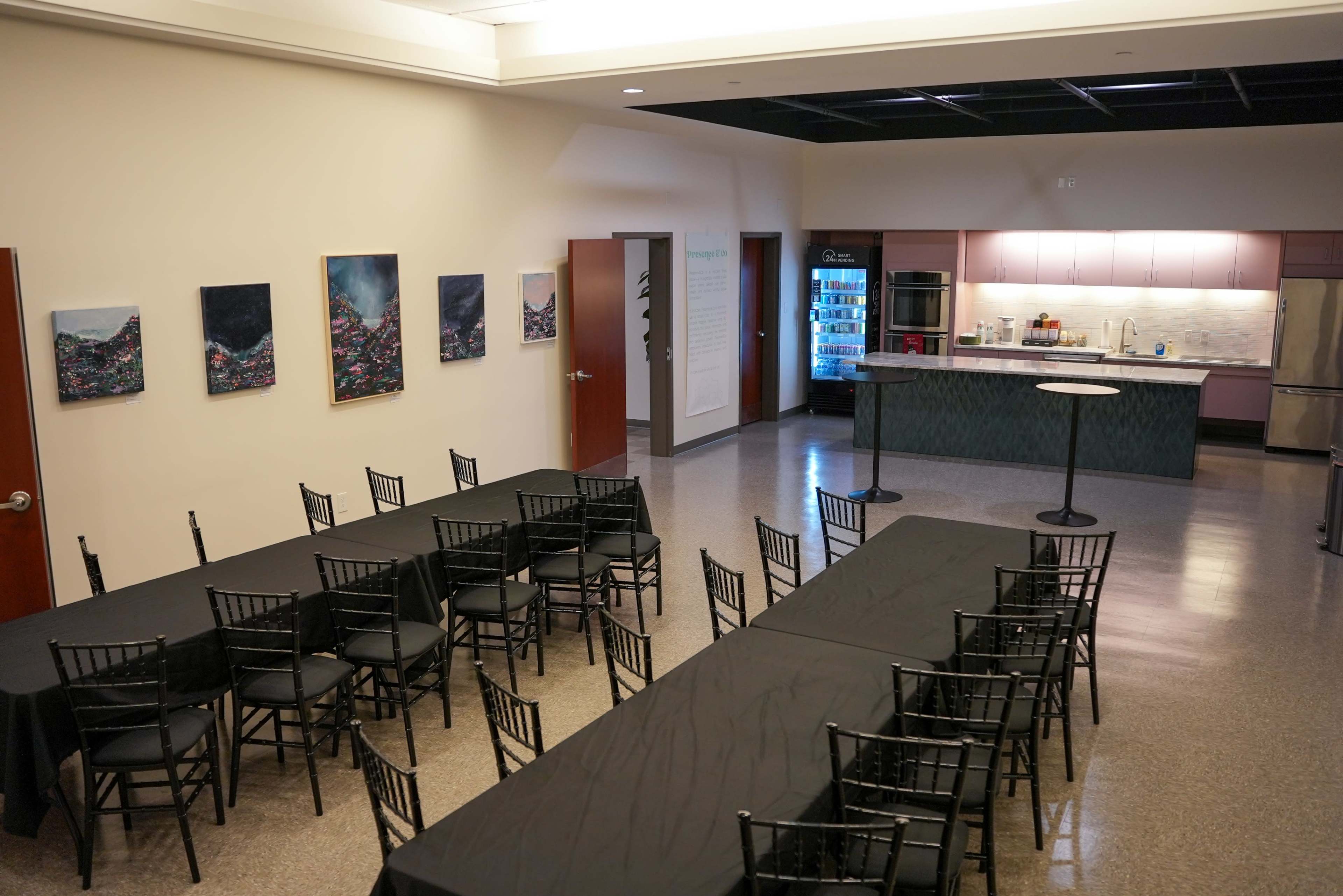 The image shows a meeting room with several black tables and chairs, along with a kitchenette area featuring a refrigerator and a wall of artwork.